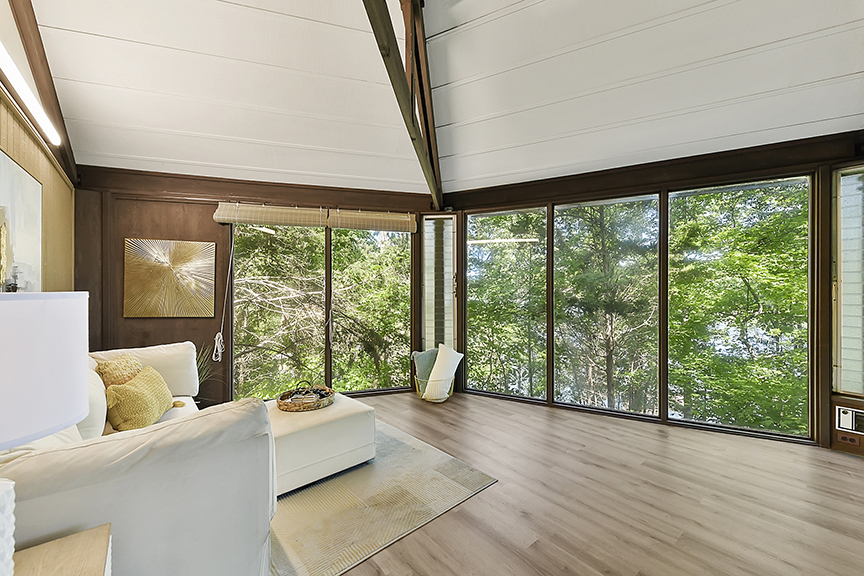 A spacious living room with large floor-to-ceiling windows offering a view of lush green trees. The room features a white sectional sofa with yellow and beige cushions, a light-colored area rug, and modern decor including a wall art piece with a sunburst design. The ceiling is high with exposed wooden beams.