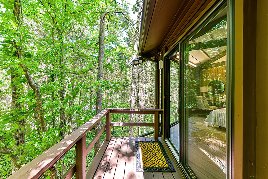 A wooden balcony with a railing, surrounded by lush green trees. The balcony is attached to a house with large glass sliding doors that reveal an interior room with a bed and bedside table.