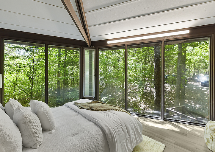 A bedroom with large floor-to-ceiling windows offering a panoramic view of a lush green forest. The room features a neatly made bed with white linens and pillows, and a light-colored rug on the wooden floor. Sunlight filters through the trees, creating a serene and natural ambiance.