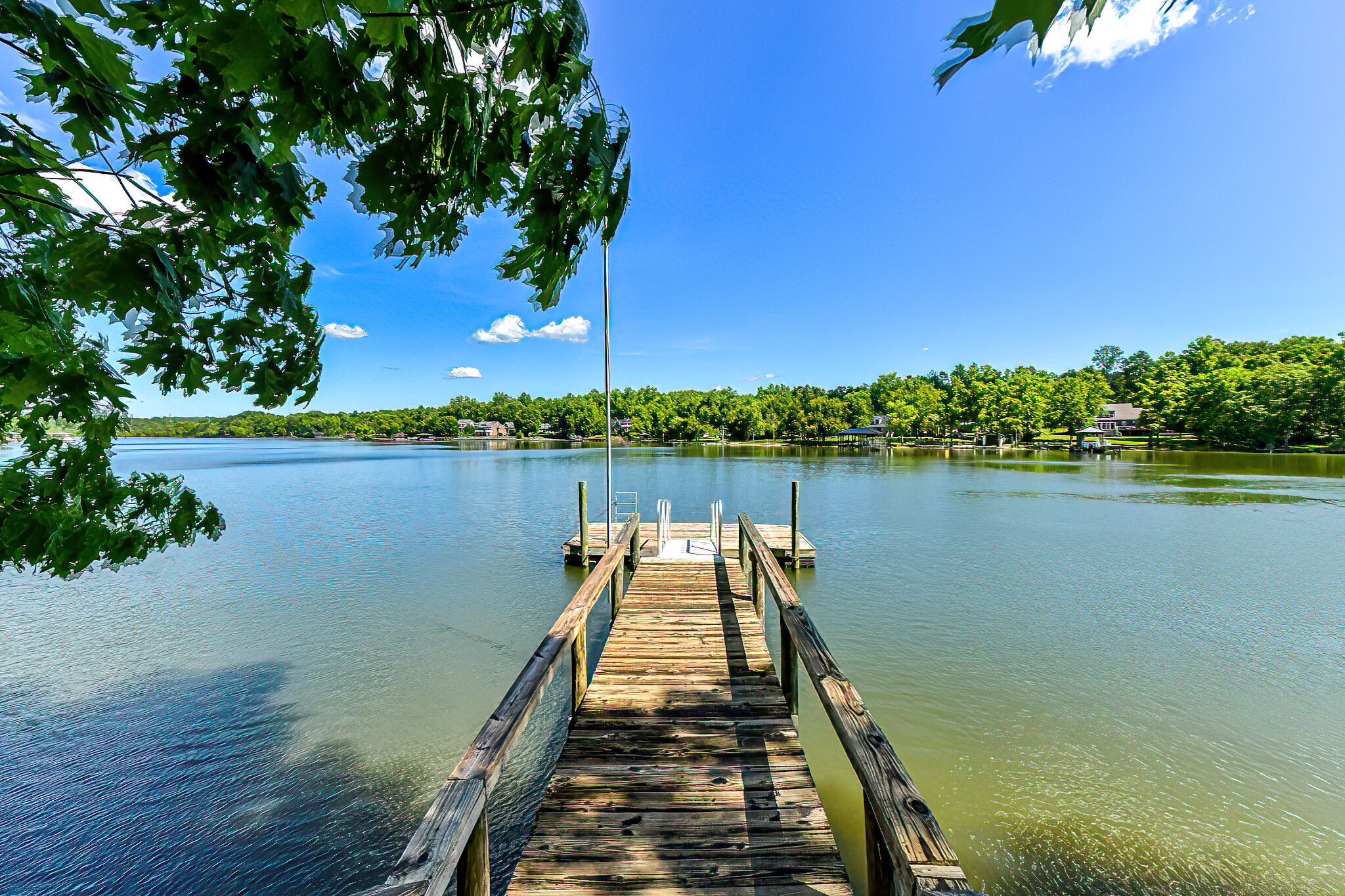 A wooden dock extends into a calm body of water surrounded by lush green trees under a clear blue sky. The dock has railings on both sides and leads to a small platform with a ladder at the end. Overhanging branches with green leaves frame the top of the image.