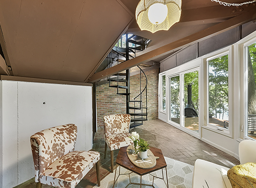 A cozy living area with two cowhide-patterned chairs, a small wooden coffee table with a plant and decorative items, large windows showing greenery outside, and a spiral staircase leading to an upper level.