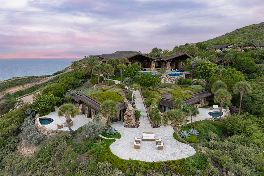 A hillside luxury property with pools, green-roofed buildings, and an ocean view under a pink sky.