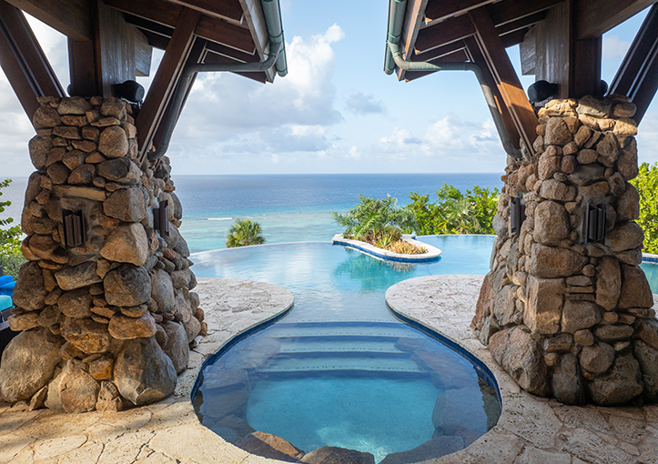 Infinity pool with a small green island, ocean view, and stone pillars under a wooden roof.