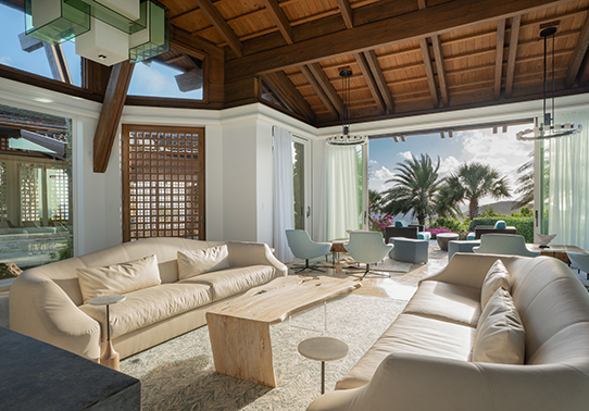 Spacious living room with beige sofas, blue chairs, large windows, and wooden ceiling beams.