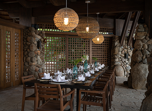 Outdoor dining table with wooden chairs, white table settings, and hanging woven lights in a rustic setting.