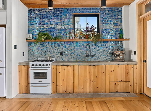 Kitchen with wooden cabinets, concrete countertop, blue tile backsplash, and plants.


