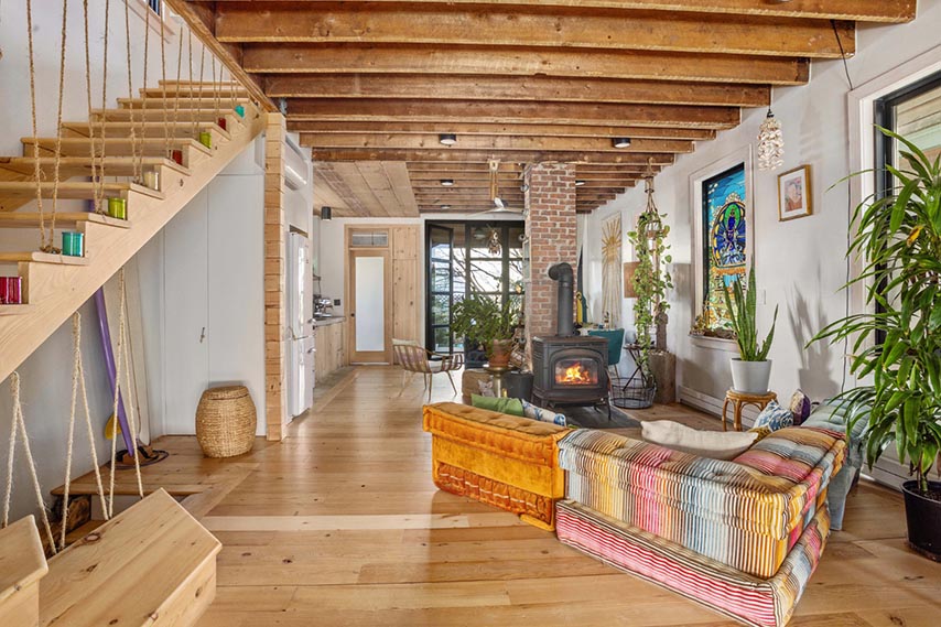 A cozy living room with wooden floors, exposed beams, a colorful couch, wood stove, plants, and large sunlit windows.