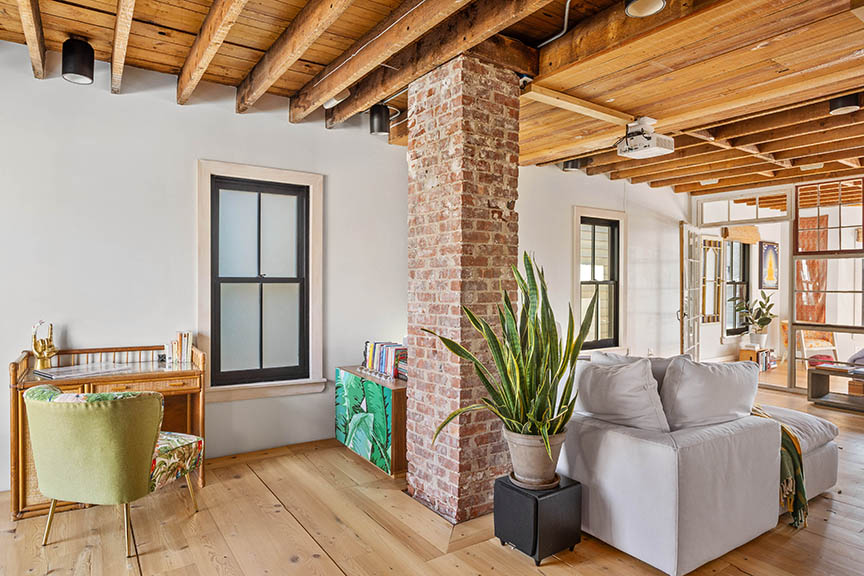 Cozy living space with wood beams, brick column, desk, sofa, and large windows.

