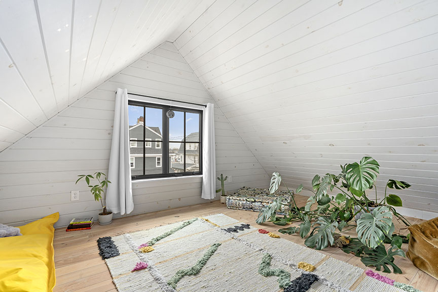 Bright attic room with white walls, plants, colorful rug, and window view.

