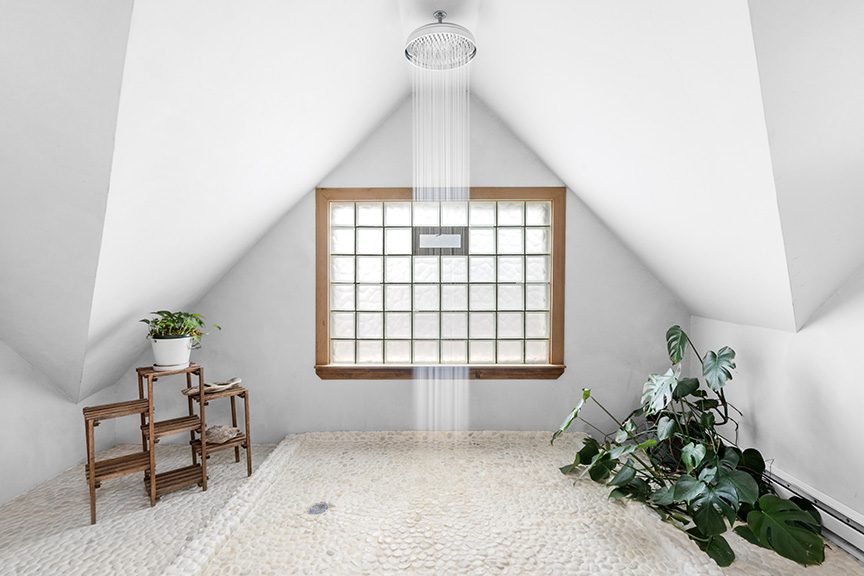 Minimalist bathroom with sloped ceiling, frosted window, rainfall shower, and plants.
