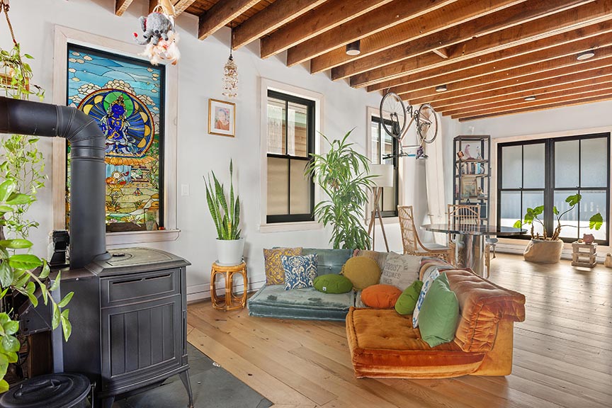 Cozy living room with wood beams, stove, plants, and stained glass window.

