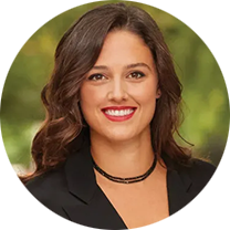 A person wearing a black blazer and a double-strand necklace, with green foliage in the blurred background.