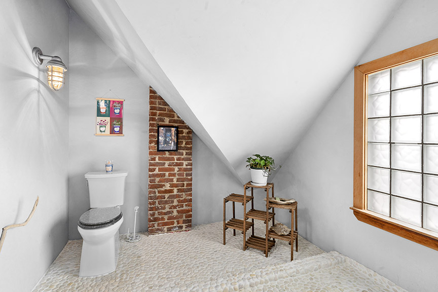 Small modern bathroom with angled ceiling, brick wall, and wooden shelf with plants.

