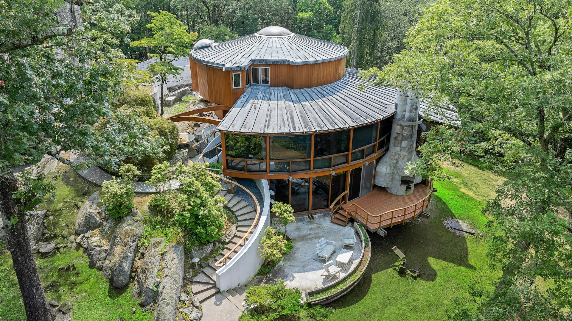 Circular modern house with glass windows and a dome roof, surrounded by trees, rocks, and grass.