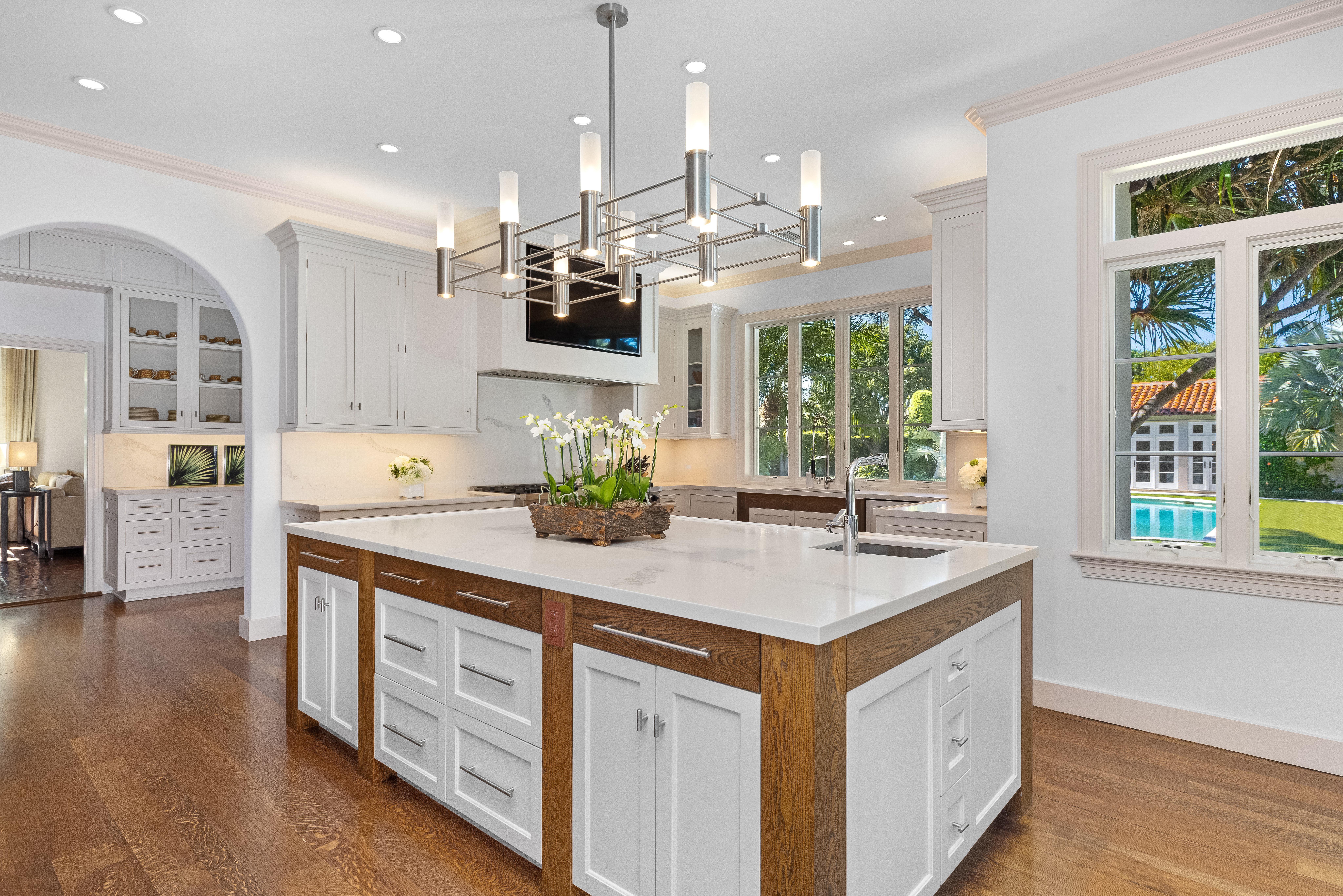 Modern luxury kitchen with large island, white cabinetry, wood accents, statement chandelier, and views of a tropical outdoor pool area.