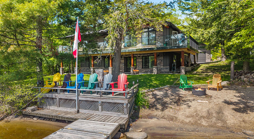 A century-old farmhouse sits next to the waterfront of Lake Muskoka in Canada's Cottage Country, with a dock and a boat.