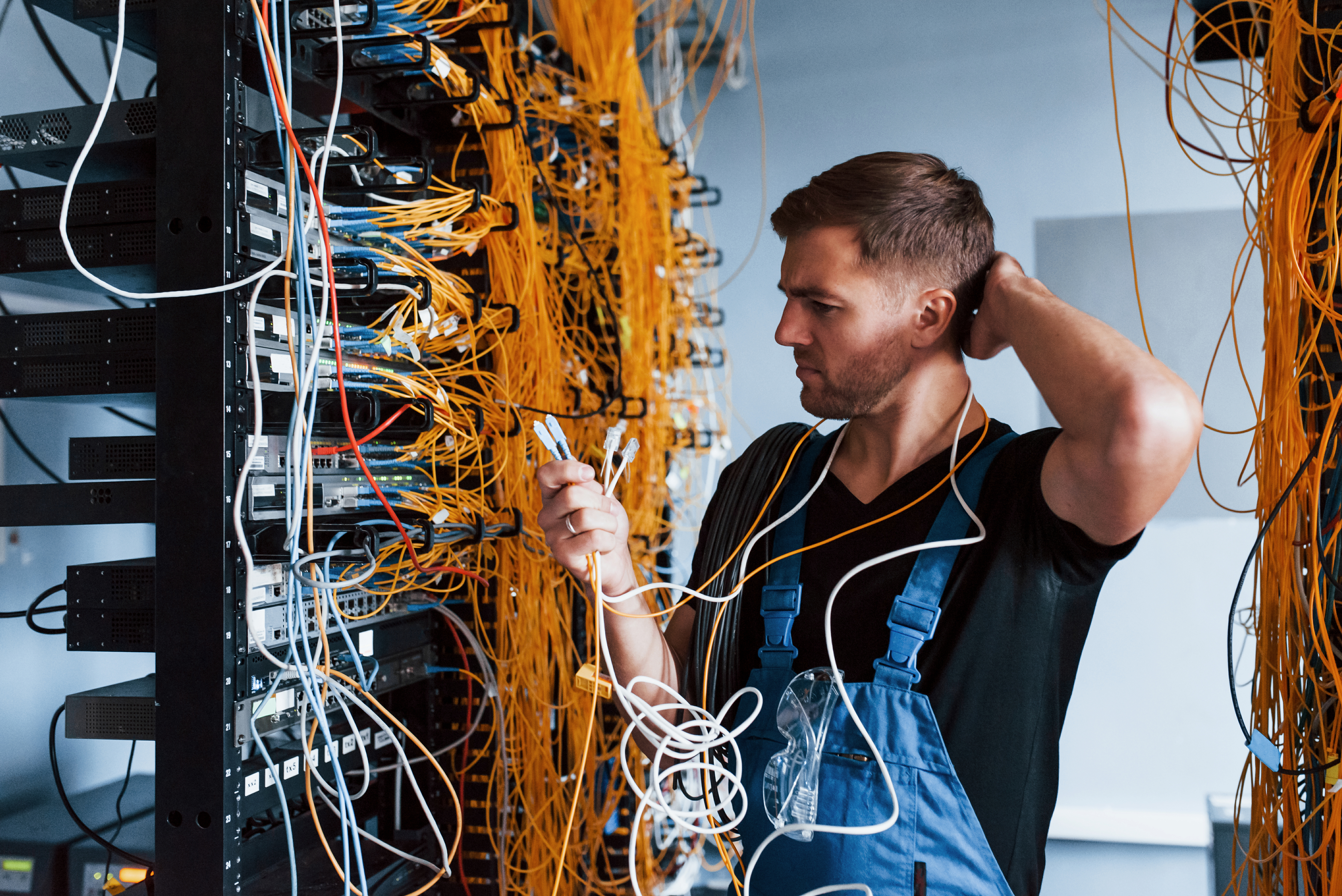 Young man in uniform feels confused and looking for a solution with internet equipment and wires in server room.