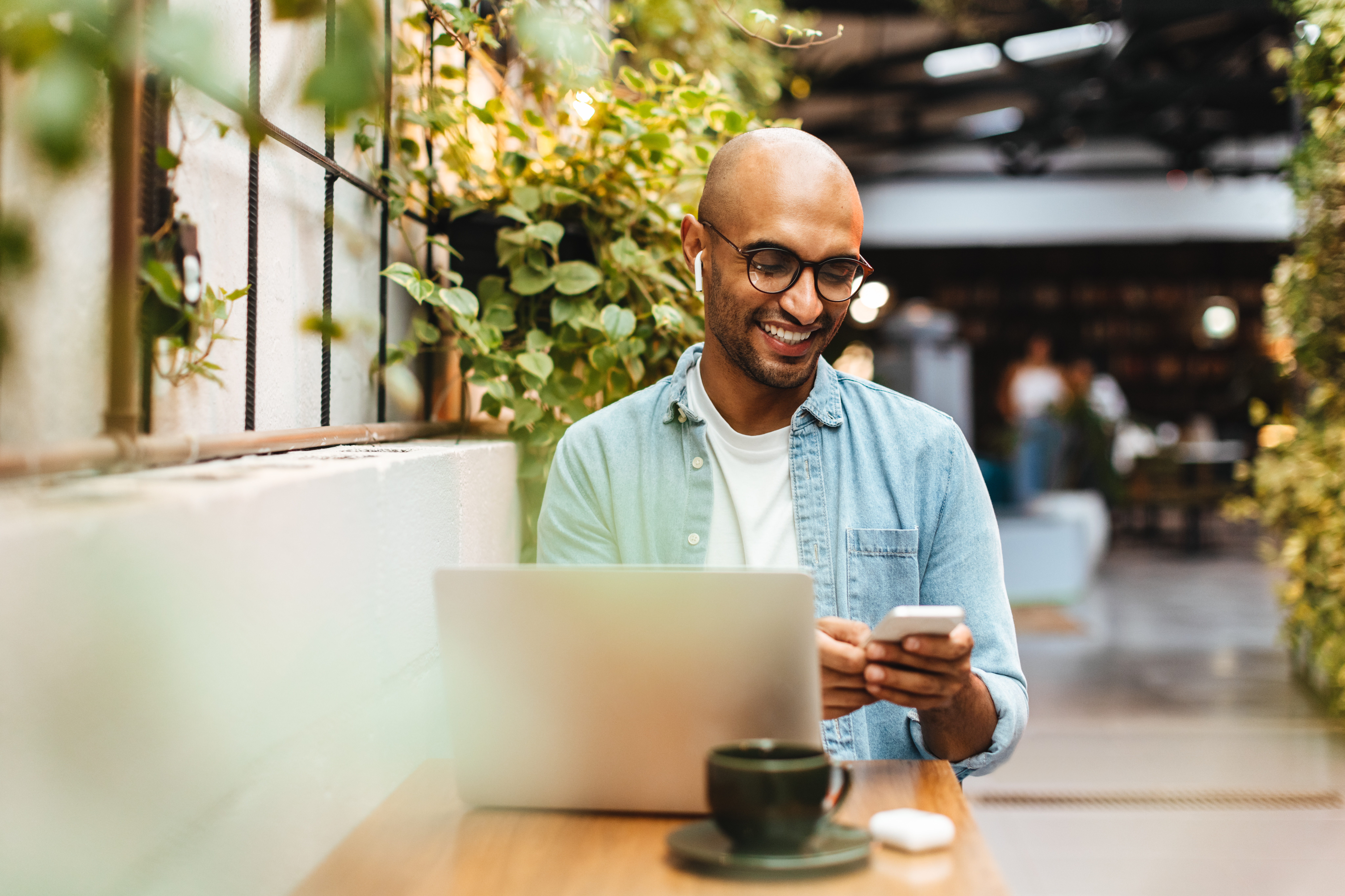 Man sits in a cafe, casually dressed and smiling while using his smartphone to play music from a streaming app. Happy young man relaxing with a laptop and a cup of coffee on the table nearby.