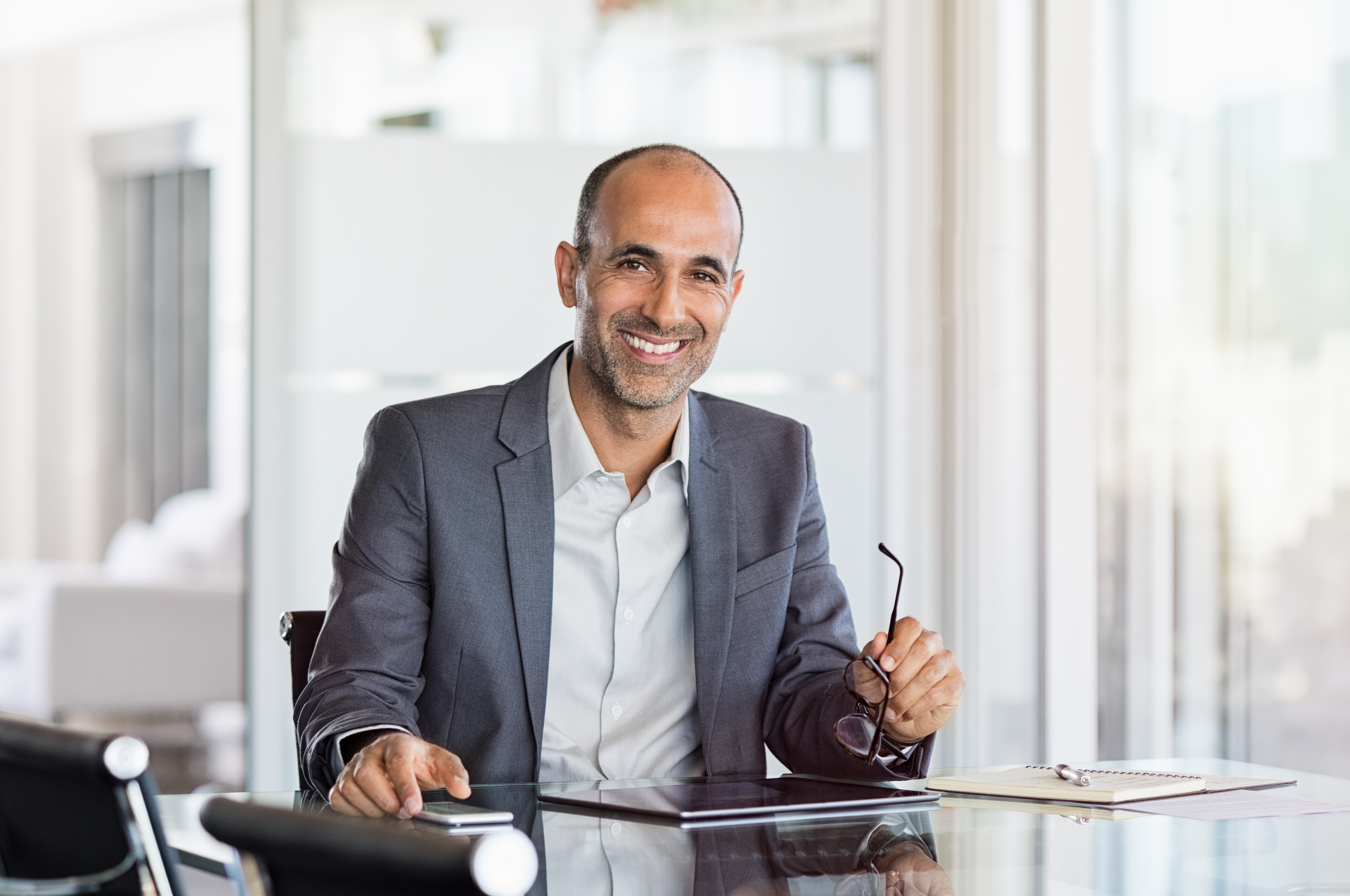 Happy mature business man holding spectacles in modern office. Successful senior businessman in formals sitting in meeting room with phone and tablet. Smiling man in suit in a elegant office.
