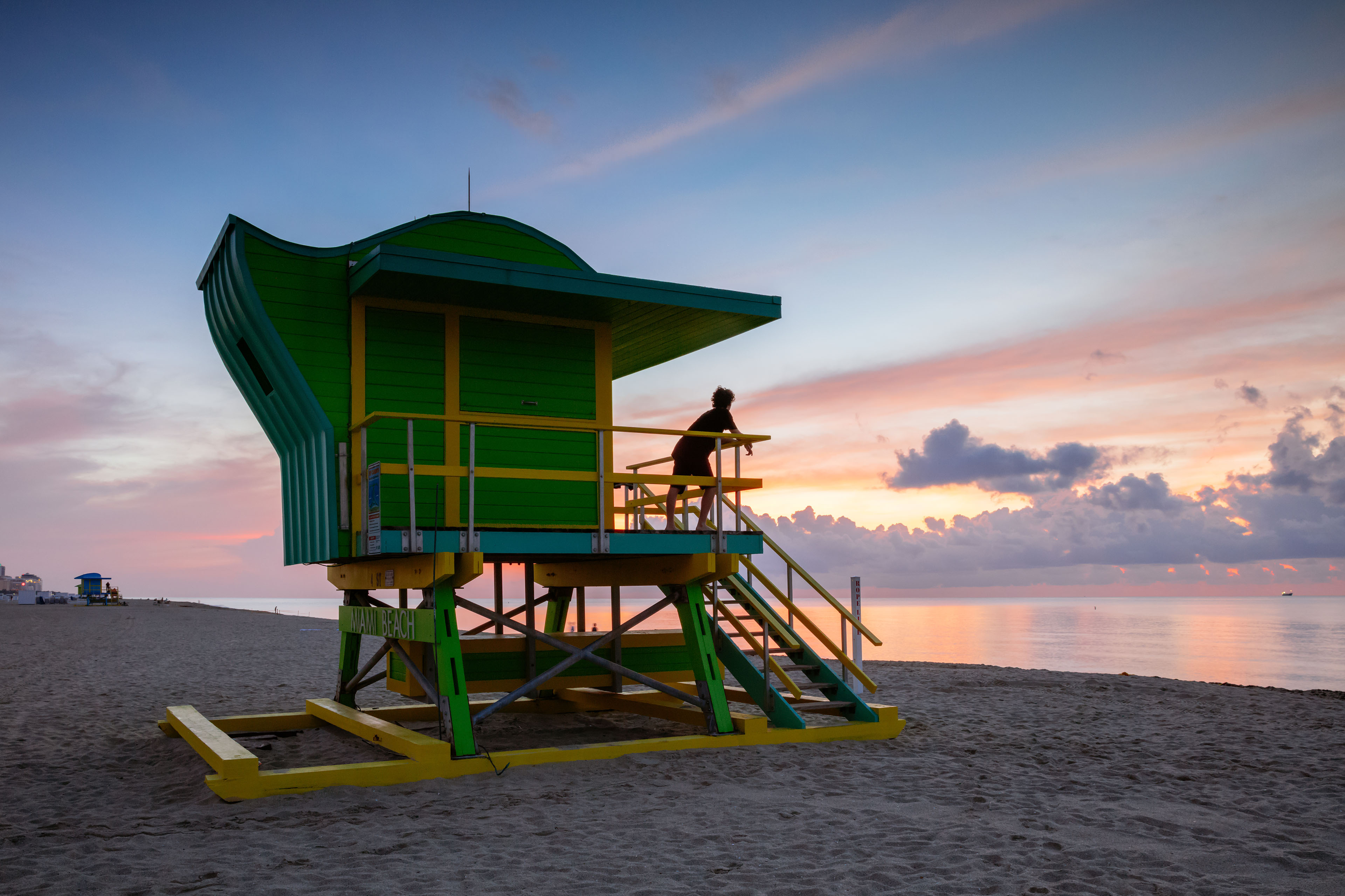 An image of a lifeguard looking out to the ocean and pink sky on the deck of a lifeguard tower
