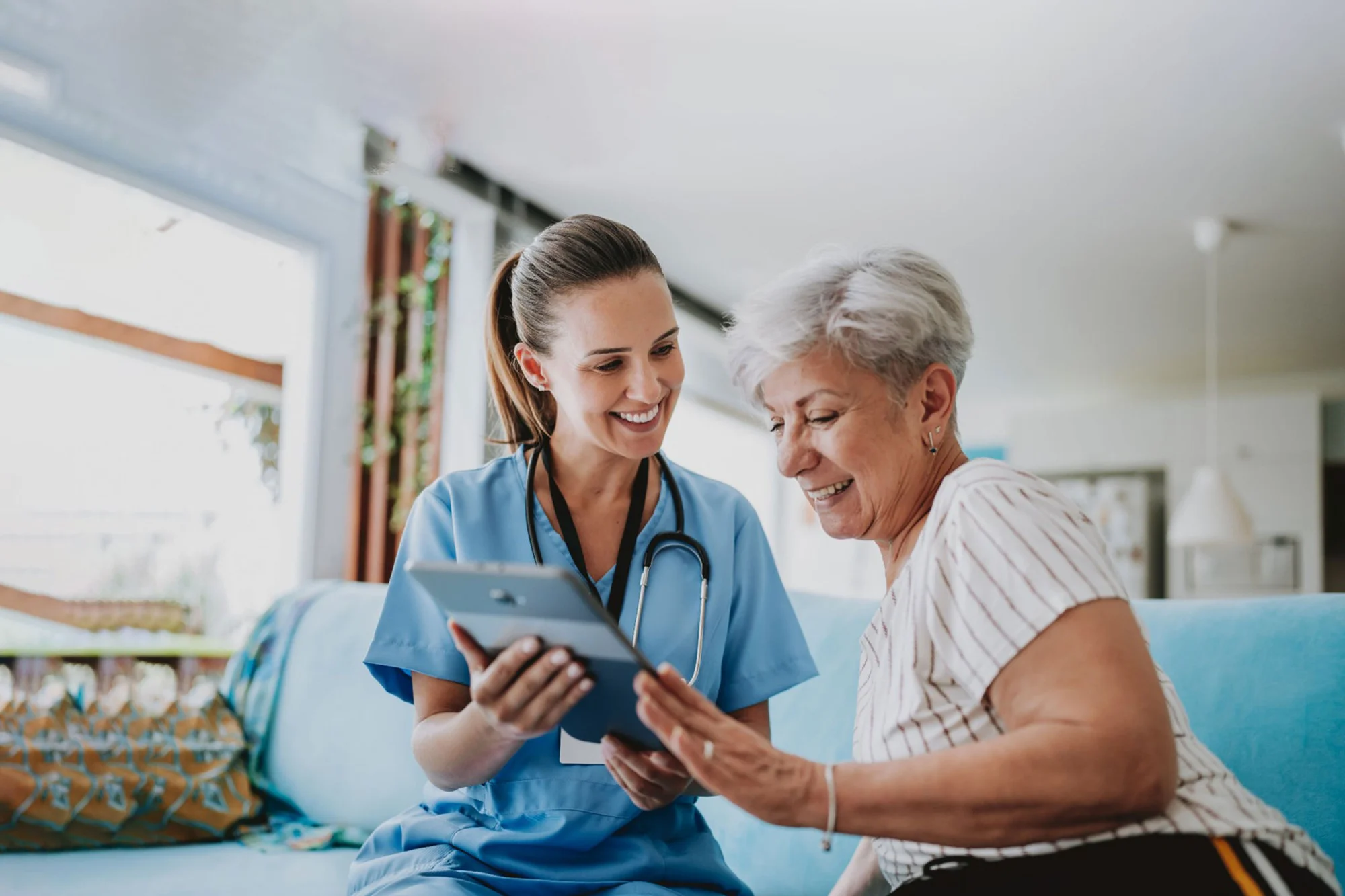 An image of a home nurse helping an elderly woman on a tablet in her living room