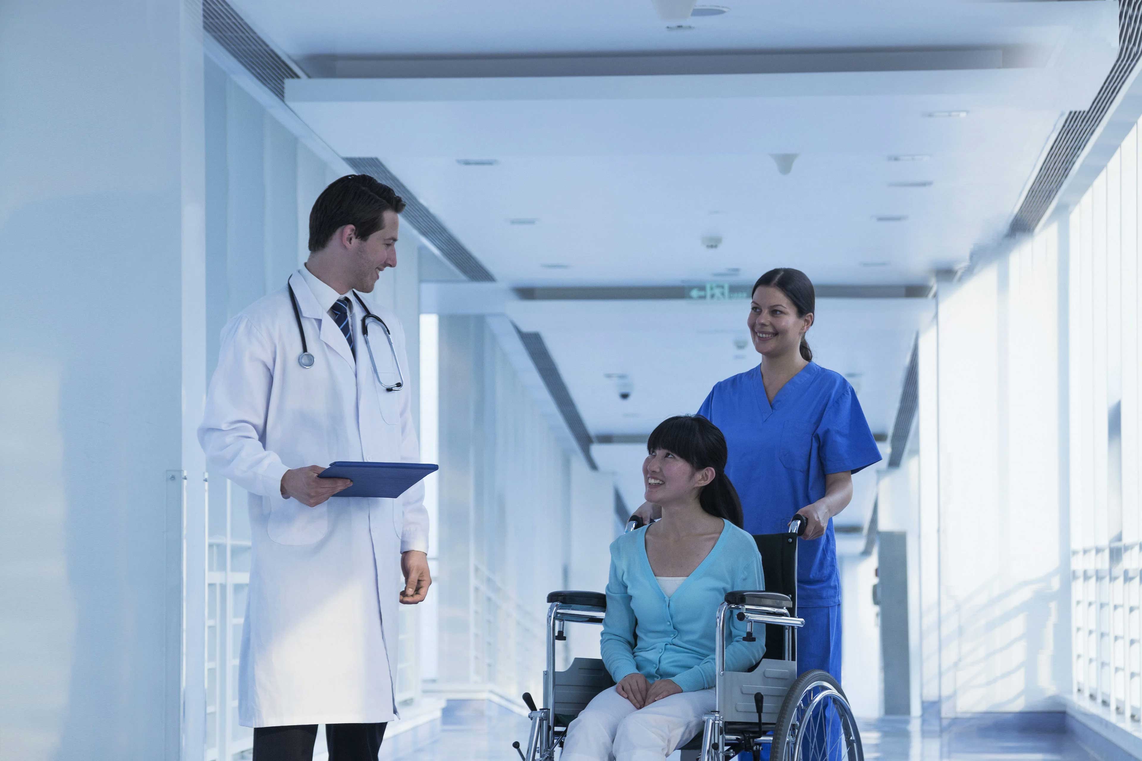 A patient being pushed by a nurse in a wheelchair talking to a doctor in a hospital hallway