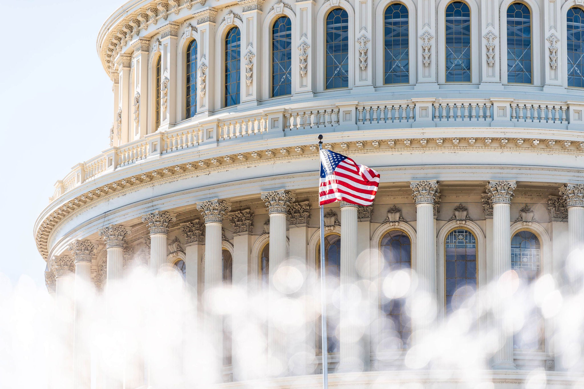 Close-up image of US Capitol dome with American flag waving and water fountain splashing in the forefront