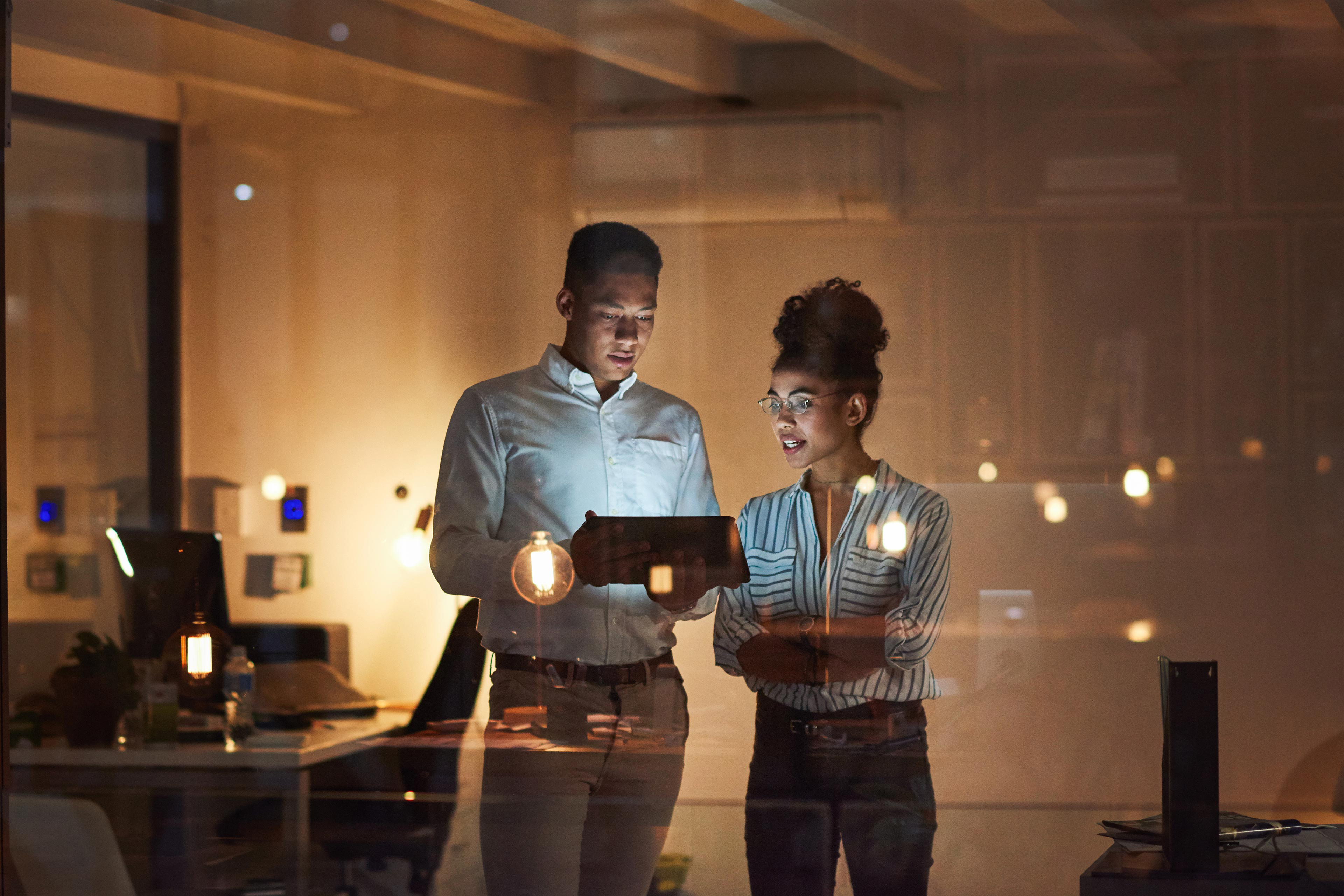 A view from outside looking into a dimly lit office with two people standing together looking at a tablet.