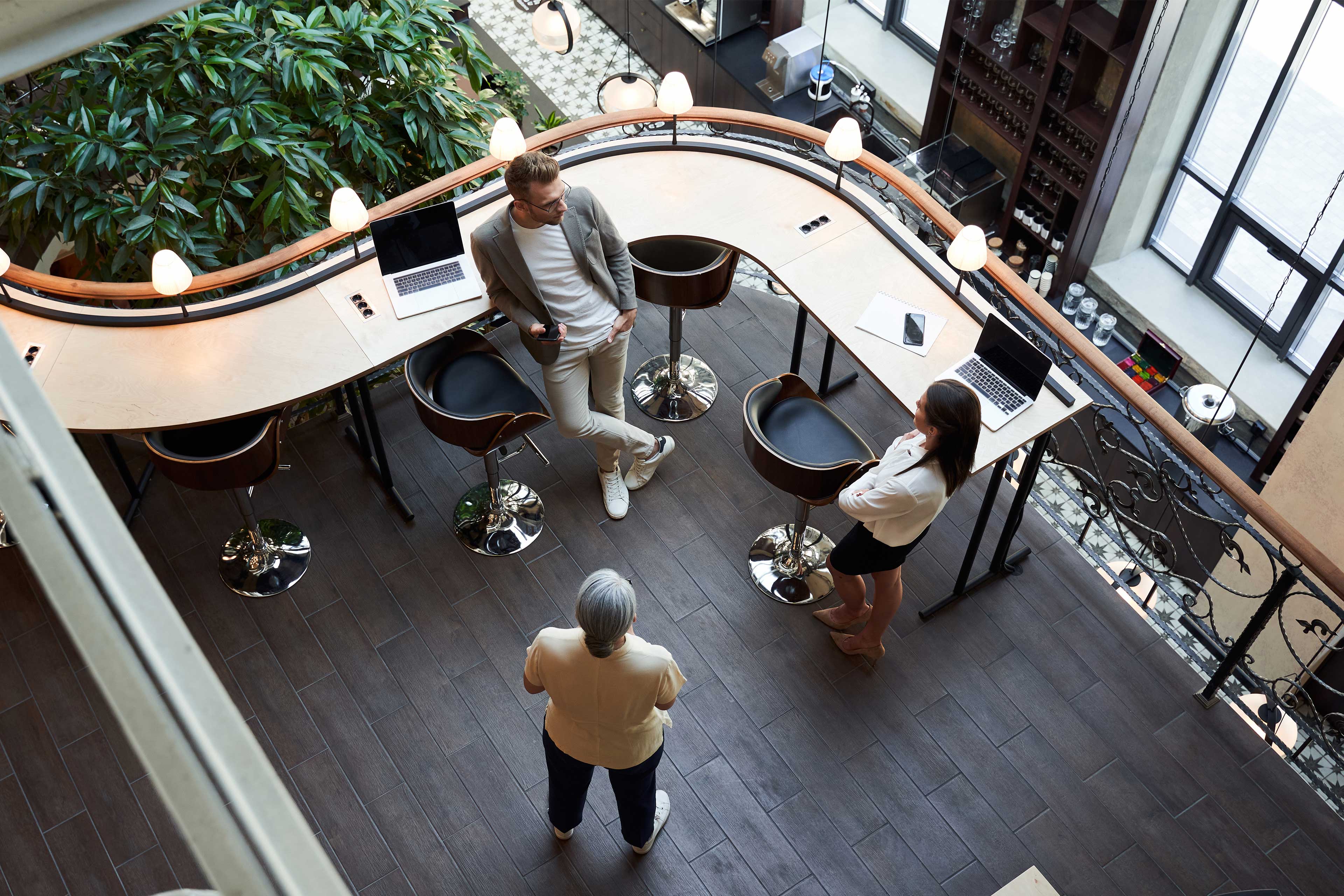 Image of three people standing and conversing in a modern workspace with laptops on a curved desk, surrounded by stylish decor and natural light.
