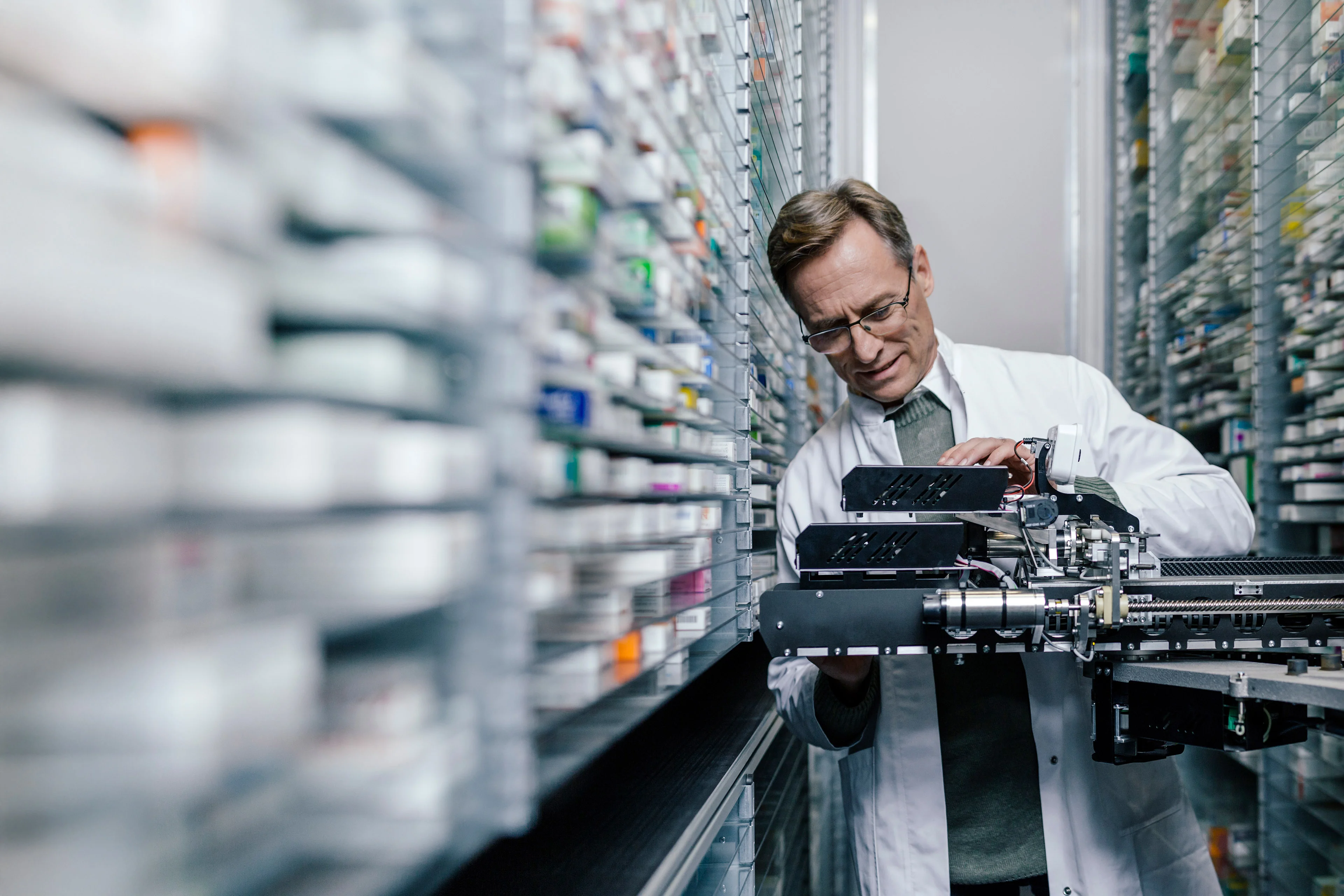 An image of a pharmacist examining a machine  in a room surrounded by shelves full of medicines.