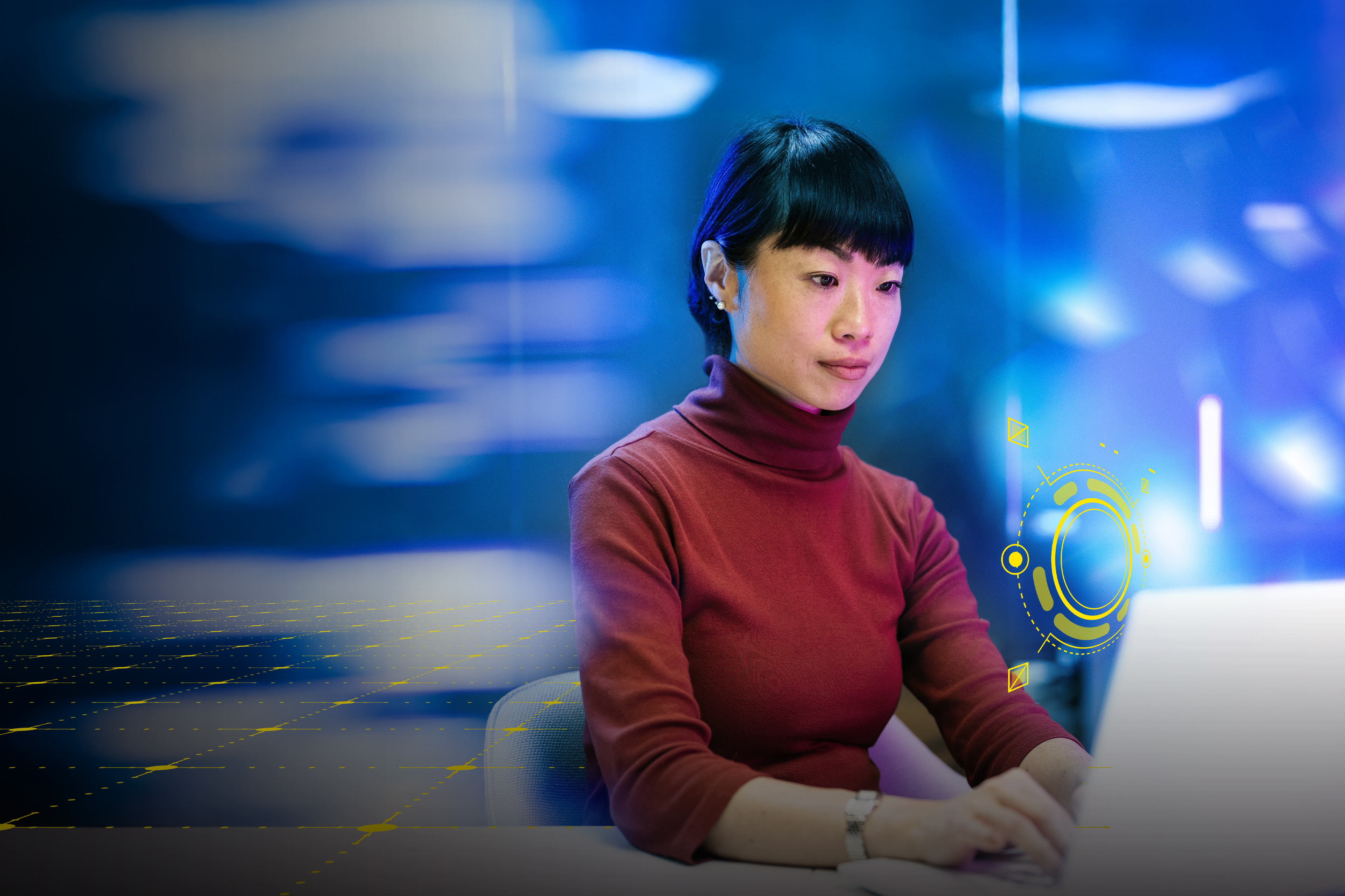 An image of a dark haired woman sitting at her desk working at her computer with a blue blurry background.