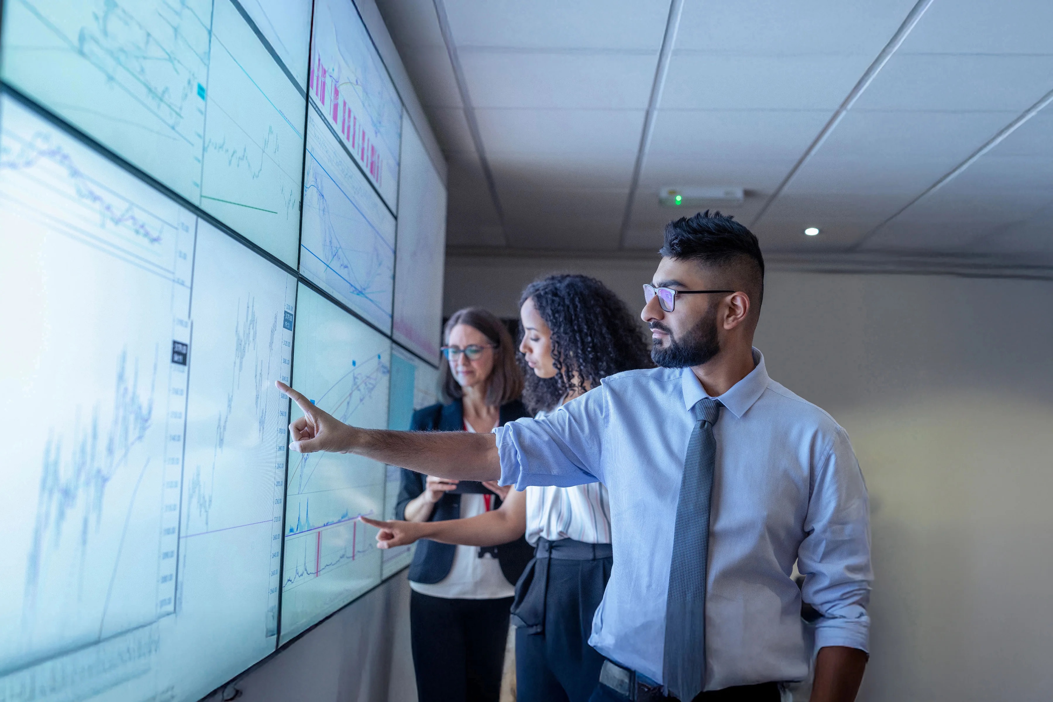 Image of three business associates in a dimmed conference room pointing and analyzing metrics data displayed on a digital screen