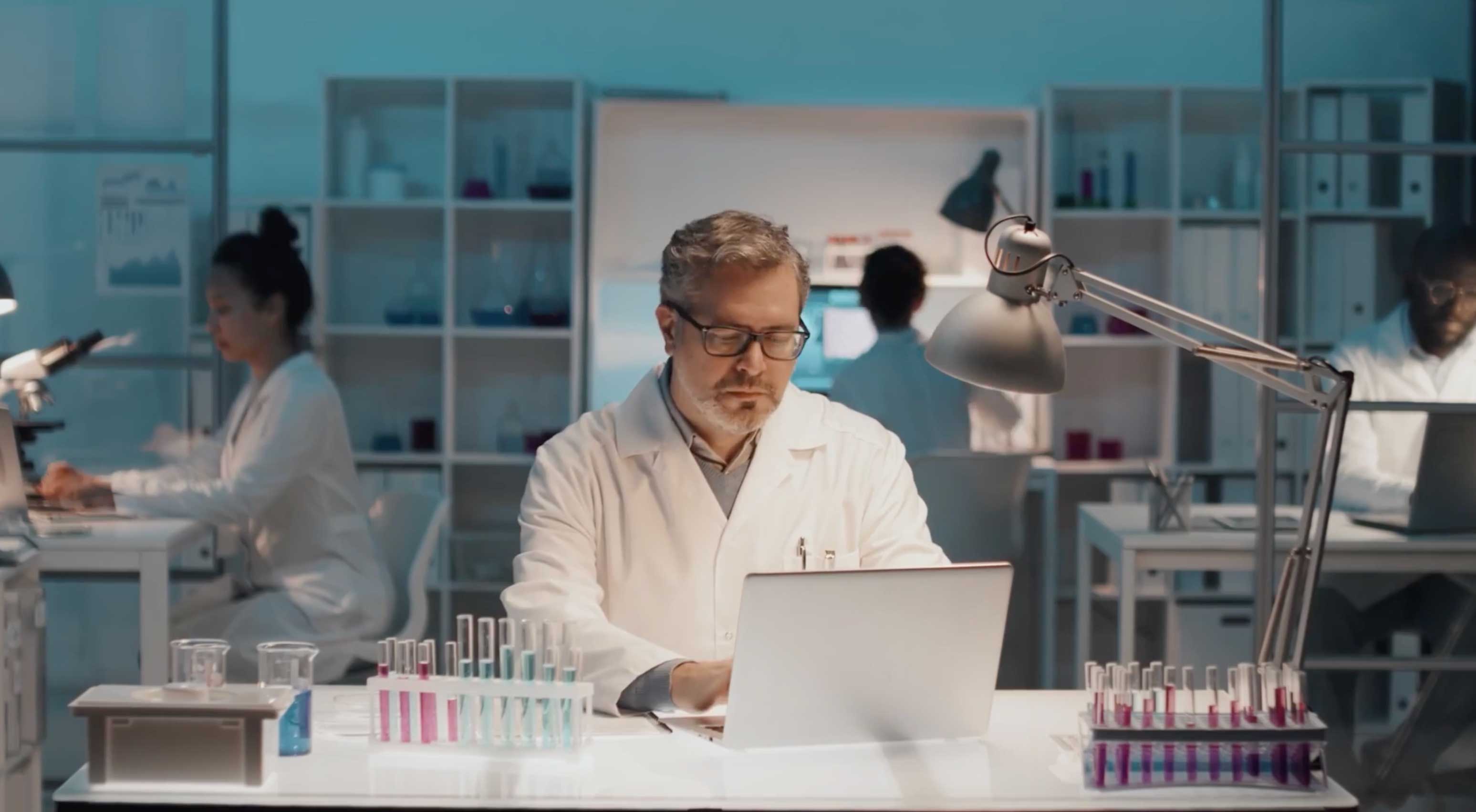 Lab tech sitting at desk working at computer surrounded by test tubes and vials