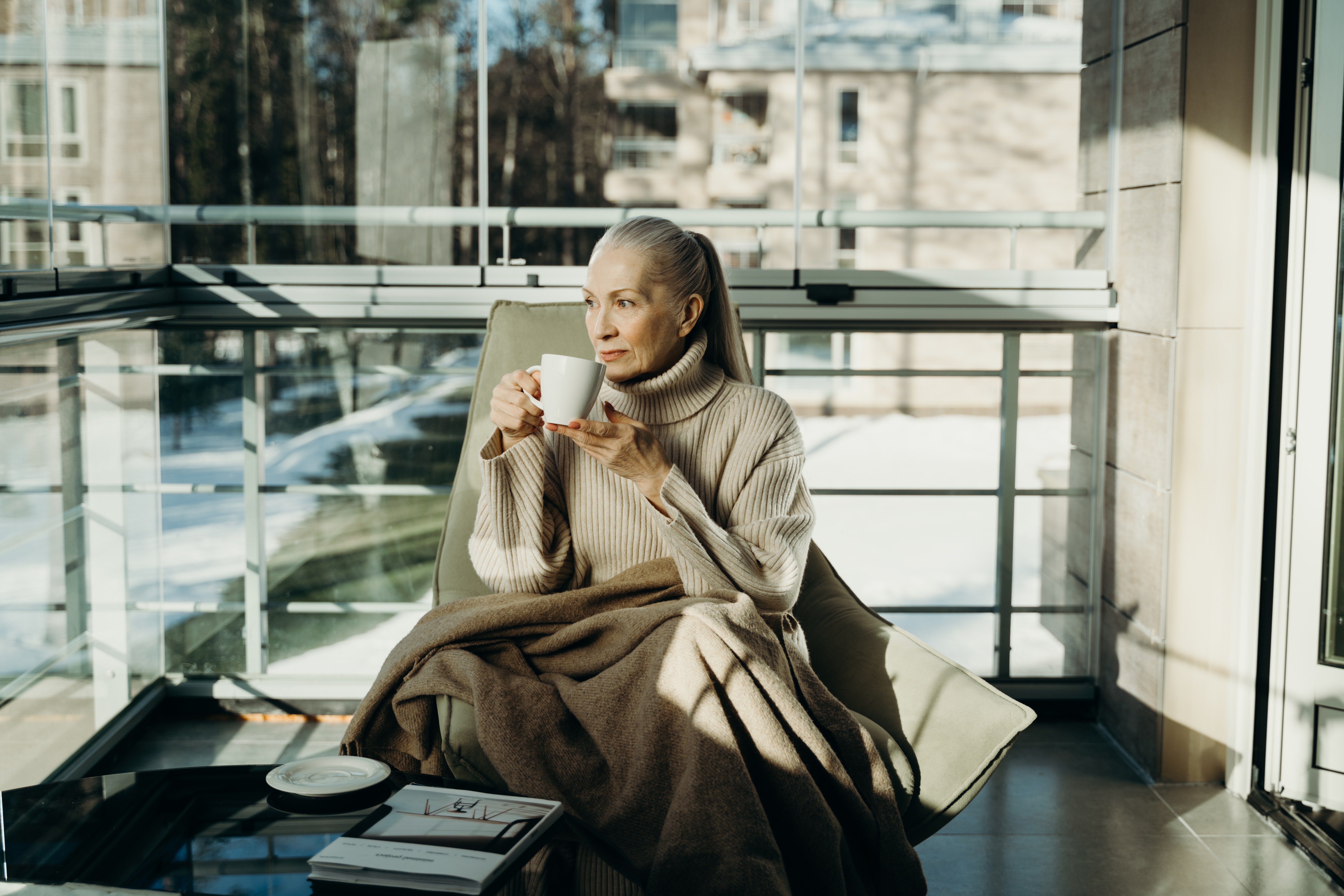 A stylish senior woman sitting down with a blanket over her legs and holding a mug