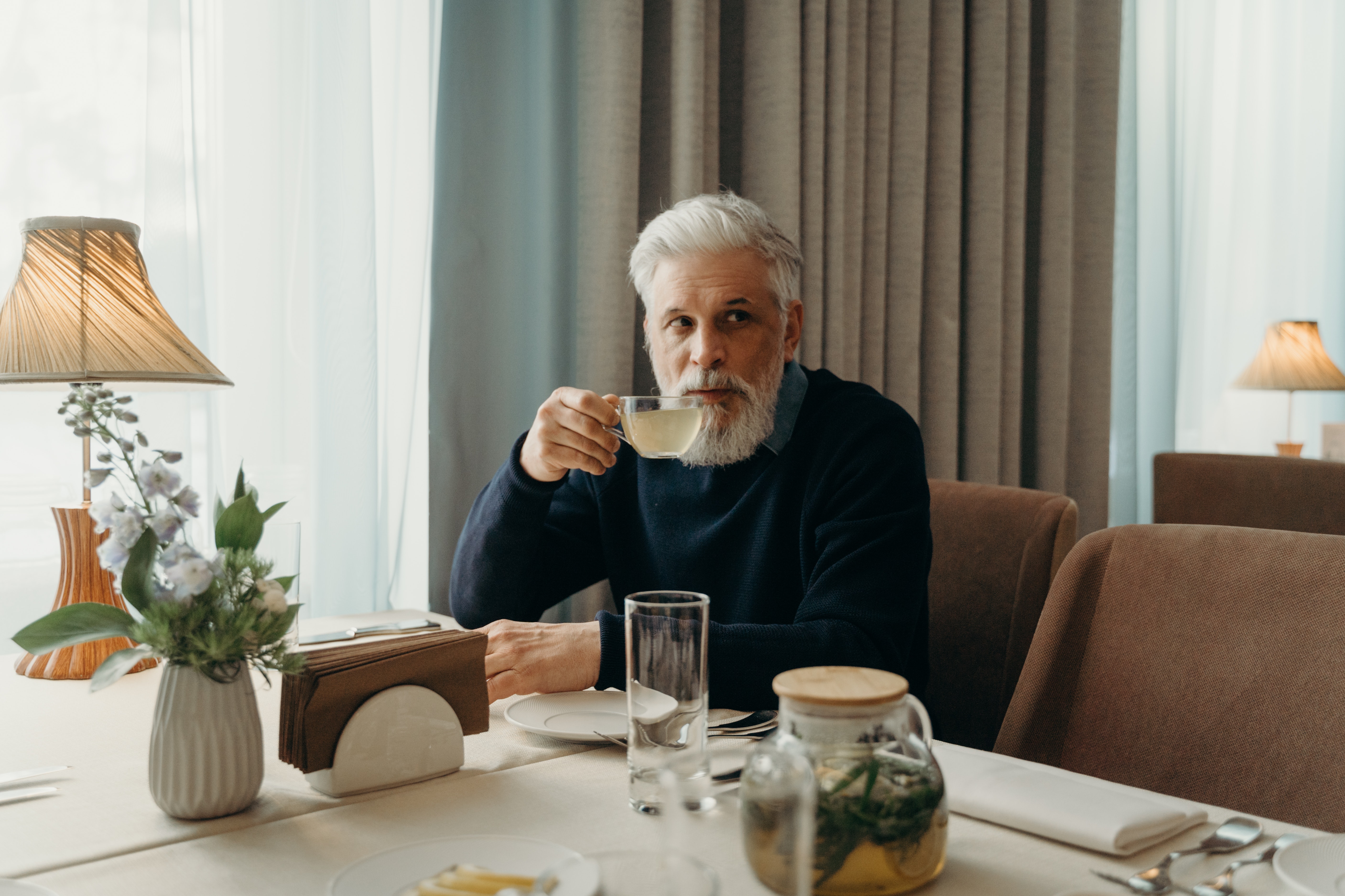A senior man with a beard holding a cup of tea in a stylish hotel room