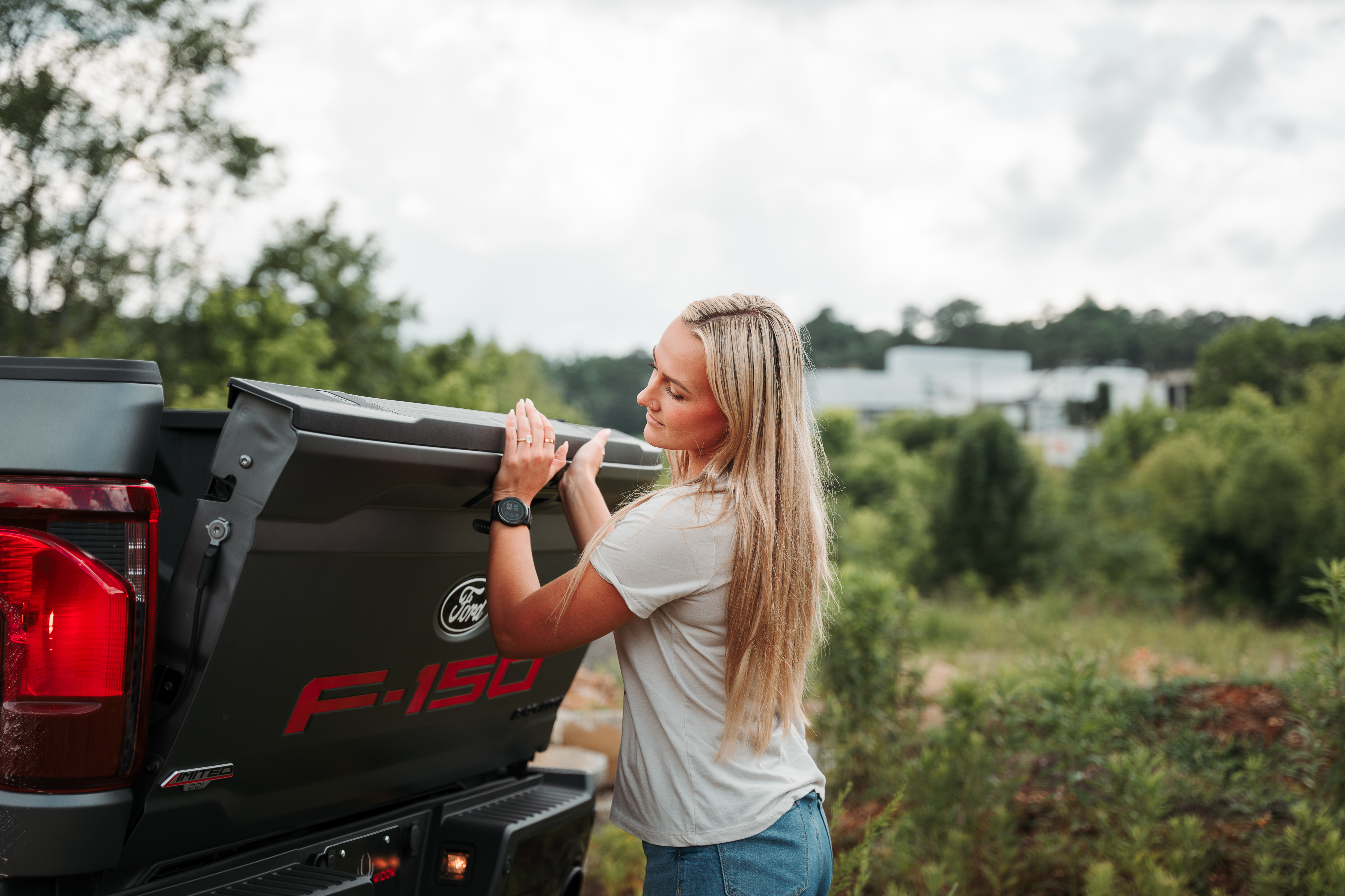 Blonde woman closing the hitch of a matte grey ford f150. Red accent colors on the badging of the black widow limited truck