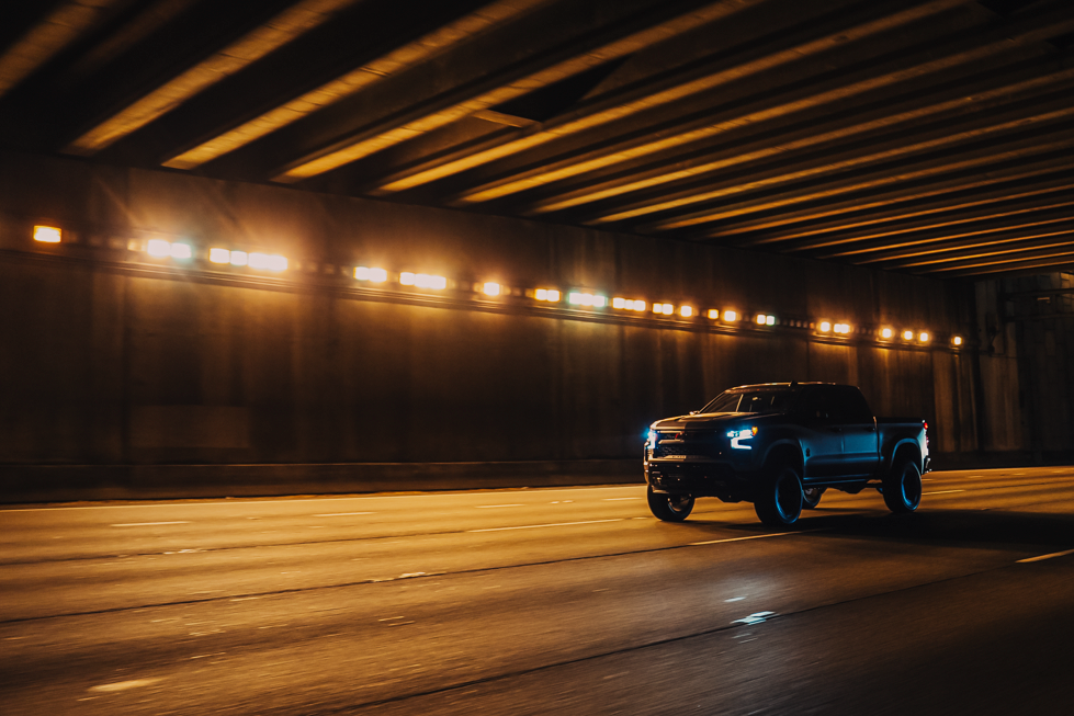 Night scene of matte grey chevrolet silverado 1500 black widow lifted truck driving under a bridge with lots of orange lights