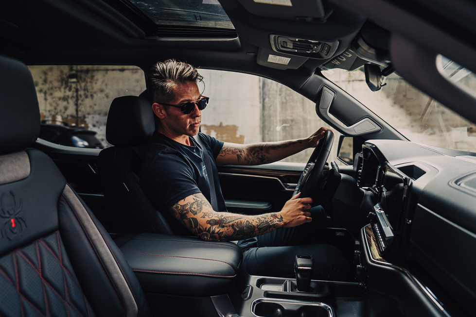 man with tattoos sitting in the driver's seat with hands on the steering wheel of a black widow lifted truck