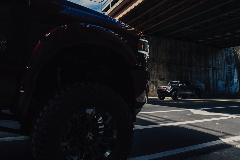 close up of a lifted black widow truck custom wheel with black widow logo a second matte grey chevrolet silverado lifted 1500 under a bridge