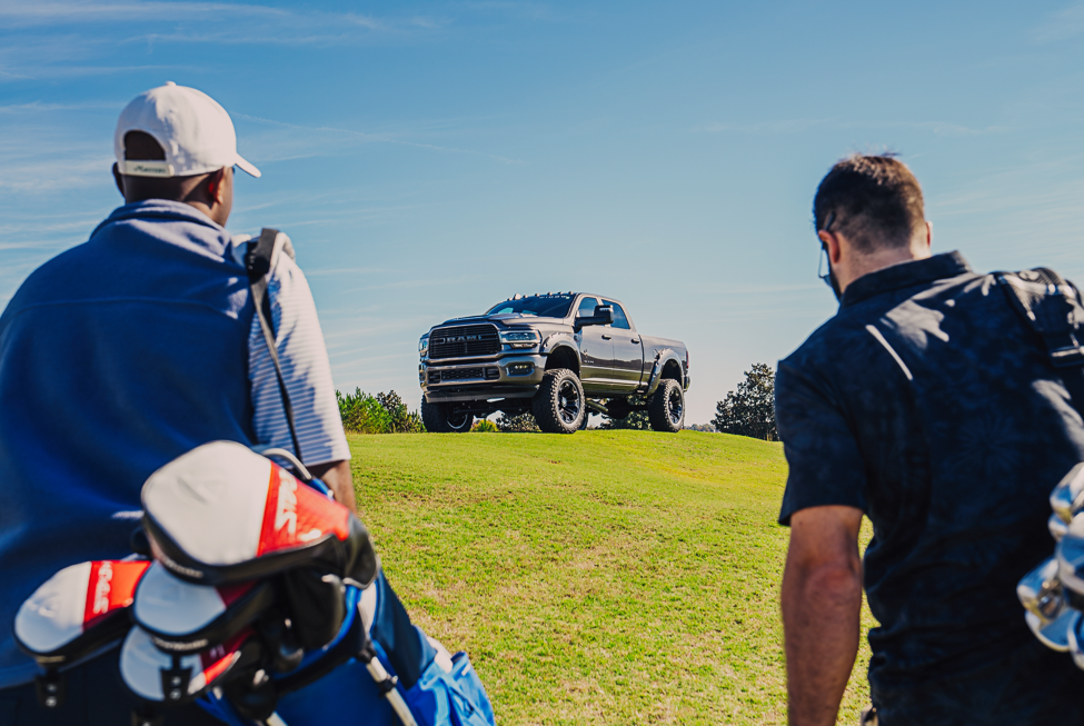 Two men carrying golf bags full of golf clubs ascent a hill towards a ram 3500 lifted black widow truck