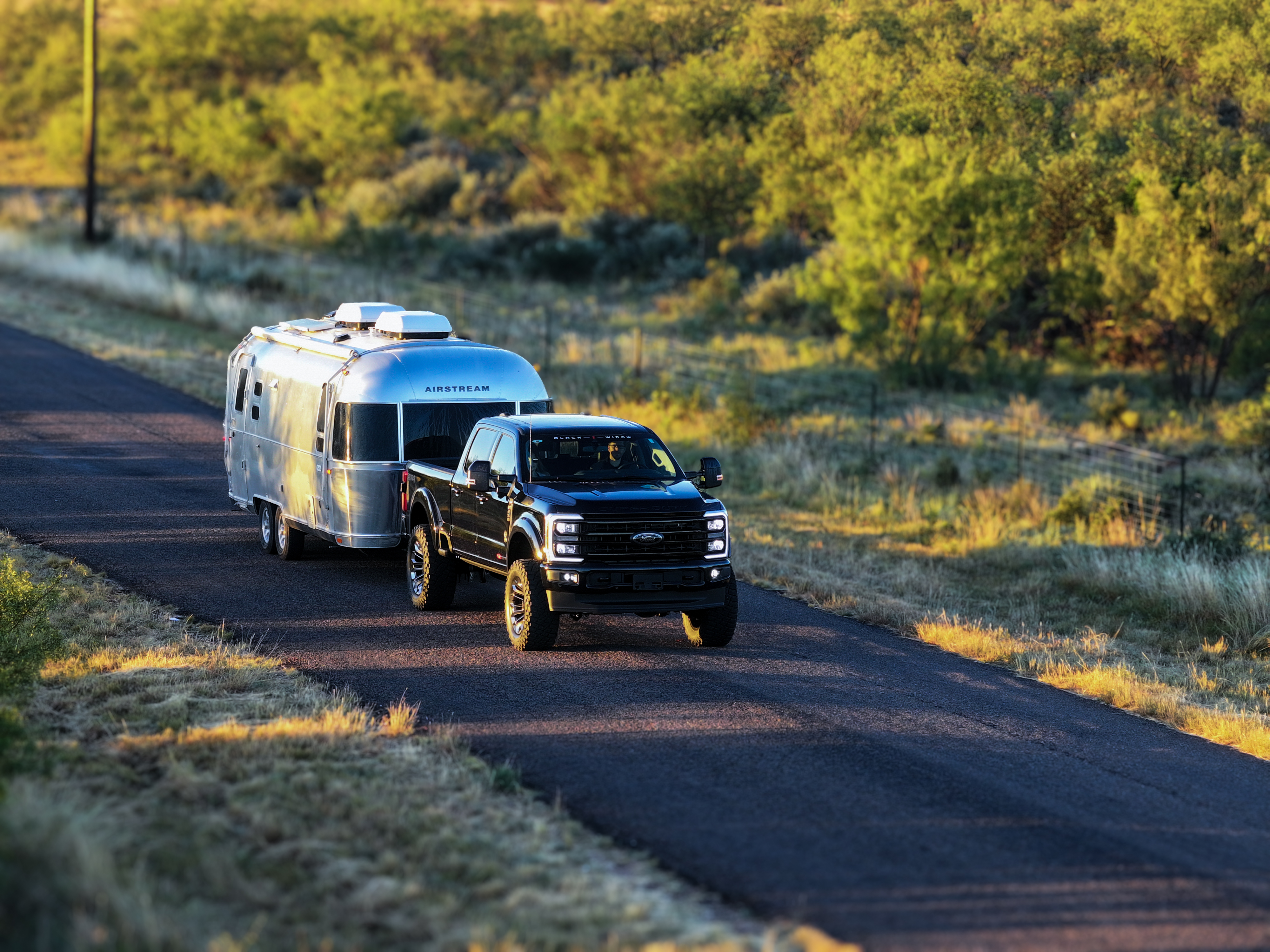 black ford superduty f250 black widow truck towing a small air stream on a two lane road at sunset