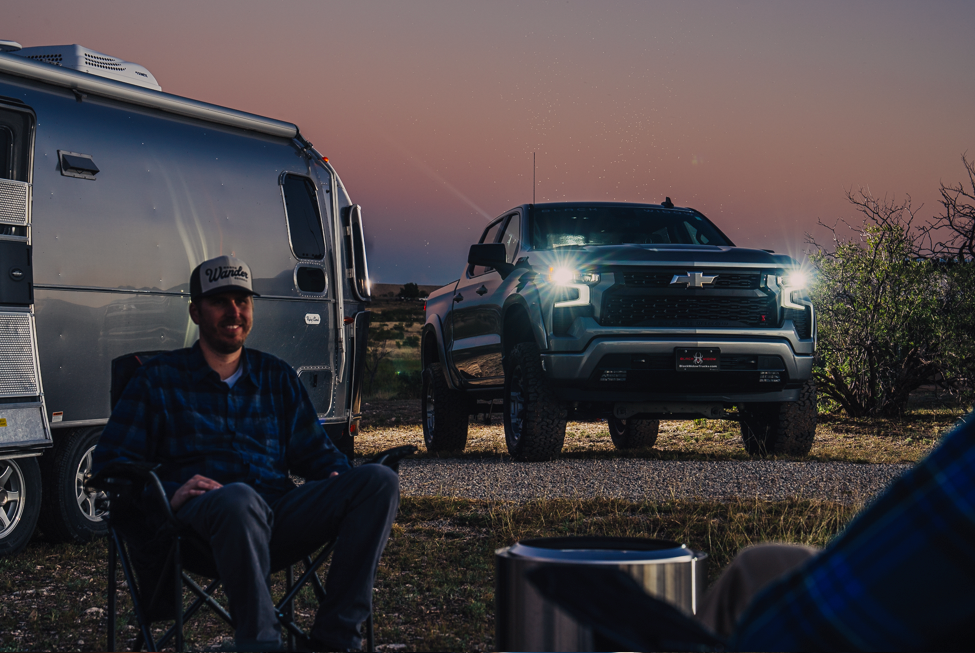 man sitting in a camping chair by a solo stove fire smiling to his friend. silver air stream behind him and the headlights of a lifted black widow silver chevrolet silverado 1500 truck shining during sunset