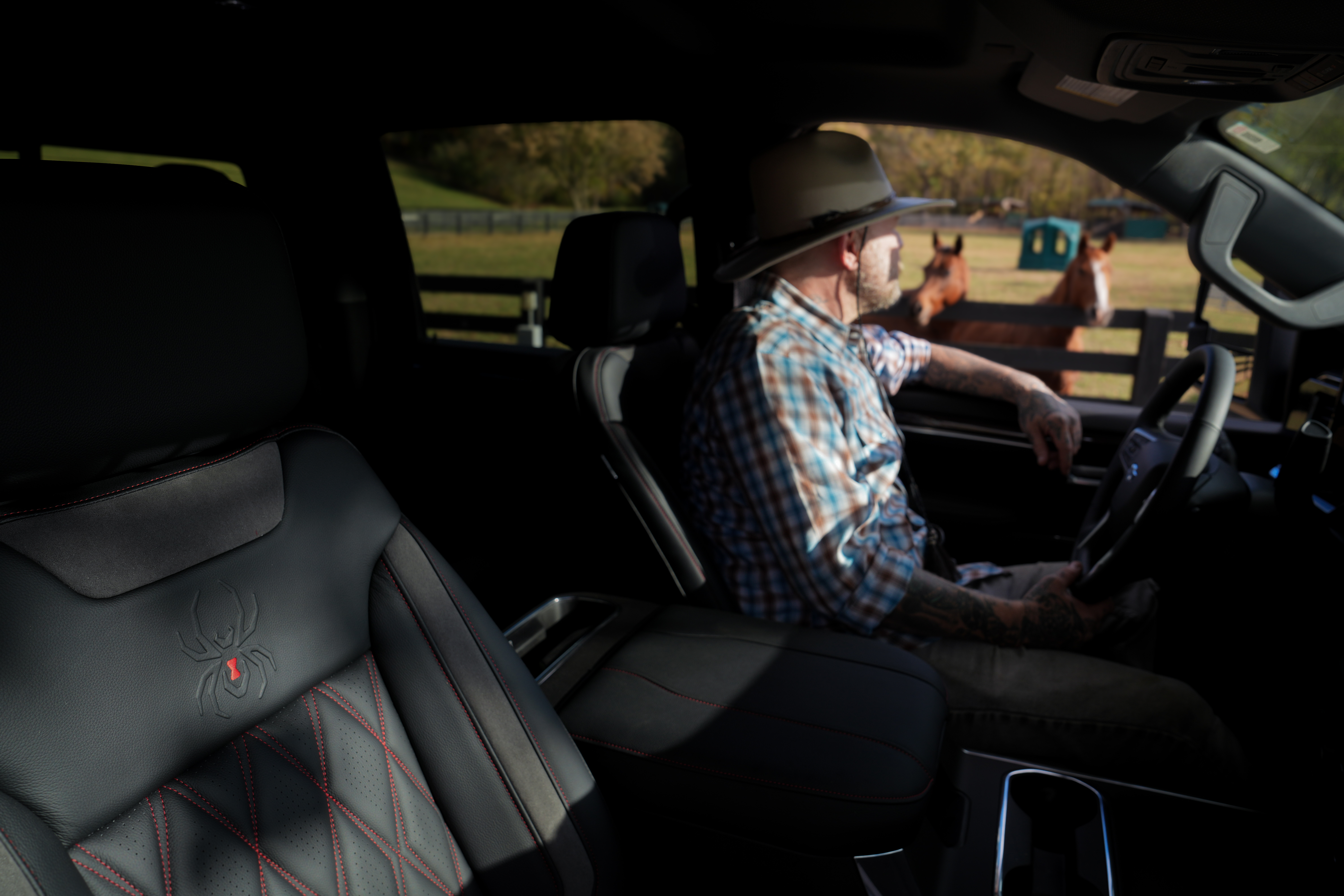 man with a stetson cowboy hat sitting in the driver's seat of a lifted black widow silverado truck with two horses in a stable