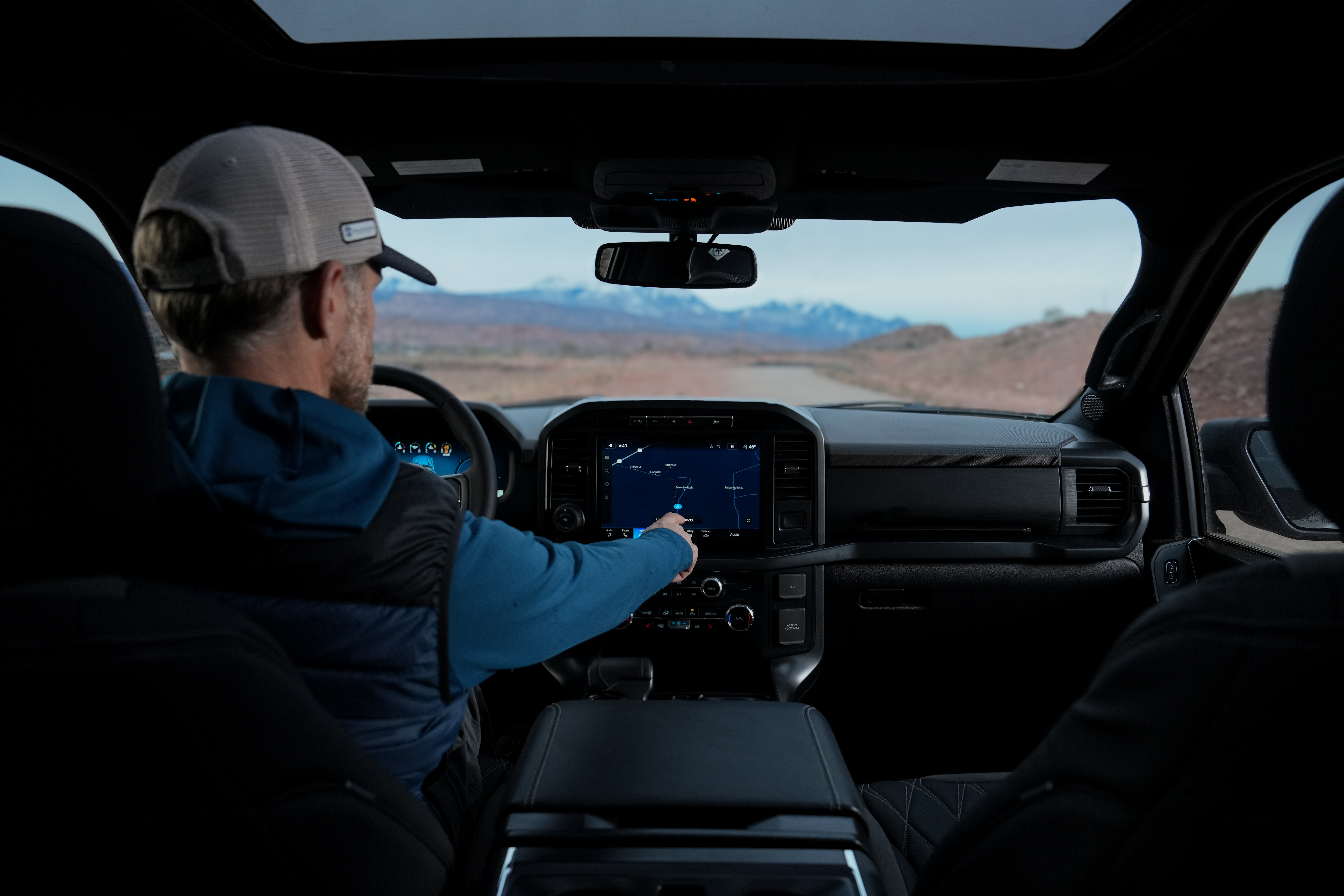 man with in a trucker hat and a adventure vest sitting in the front seat of a lifted black widow truck touching the map on his dashboard screen in the moab desert
