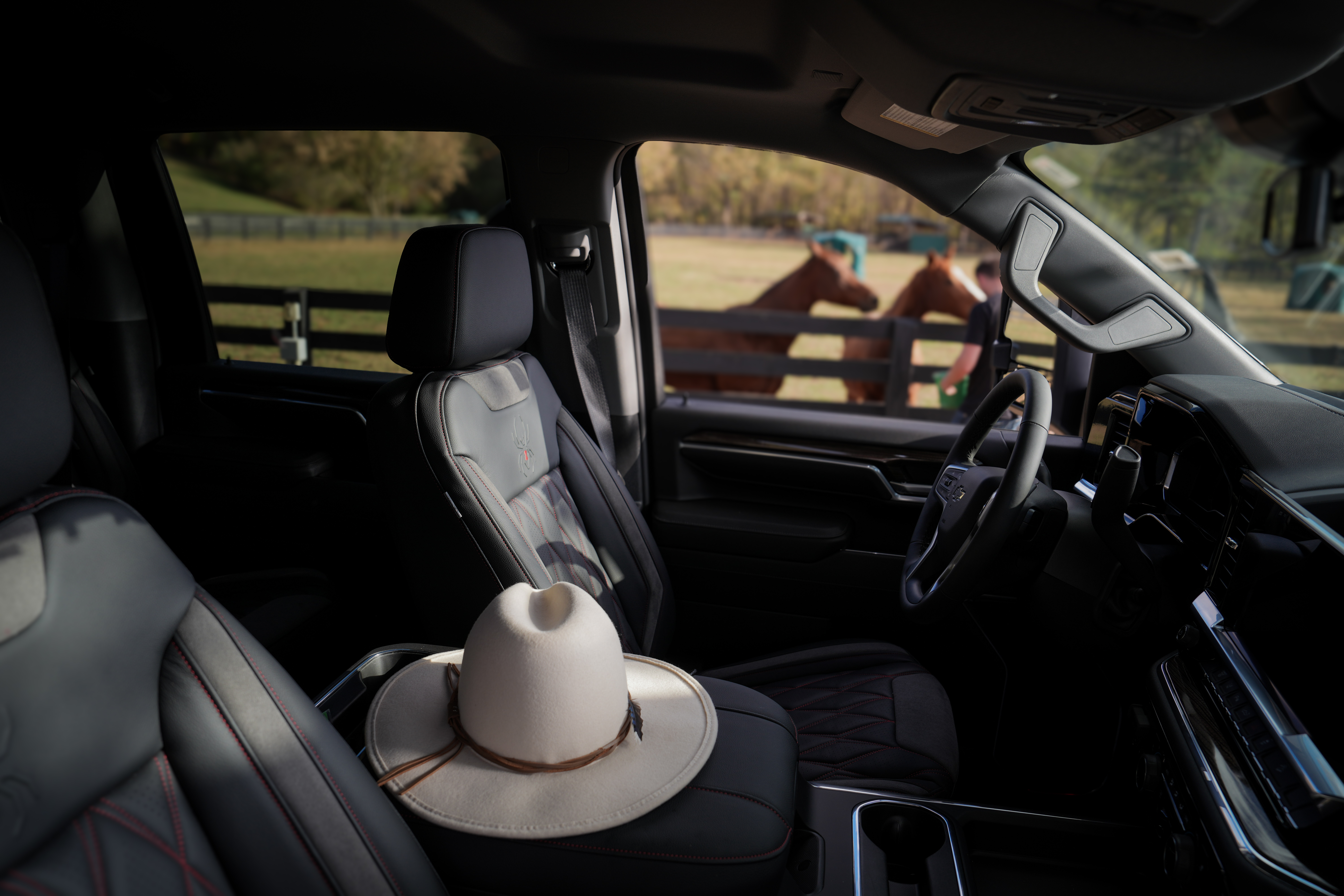 Interior view of a lifted black widow silverado lifted truck 1500. a stetson cowboy hat is sat on the center console. two horses eating are seen though the tinted windows