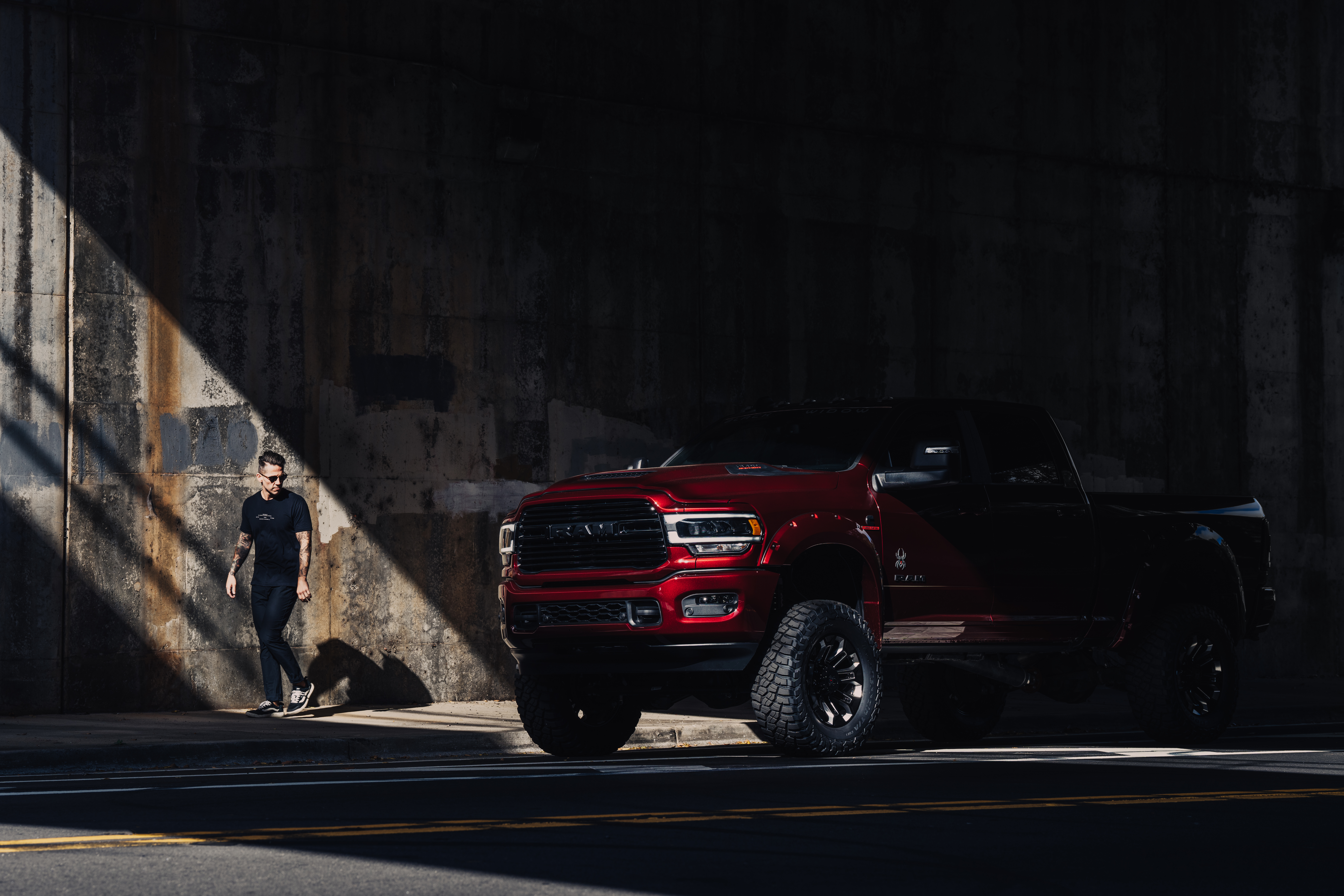 shiny red black widow lifted ram 1500 truck under a bridge with a shadow and a tattooed man in sunglasses