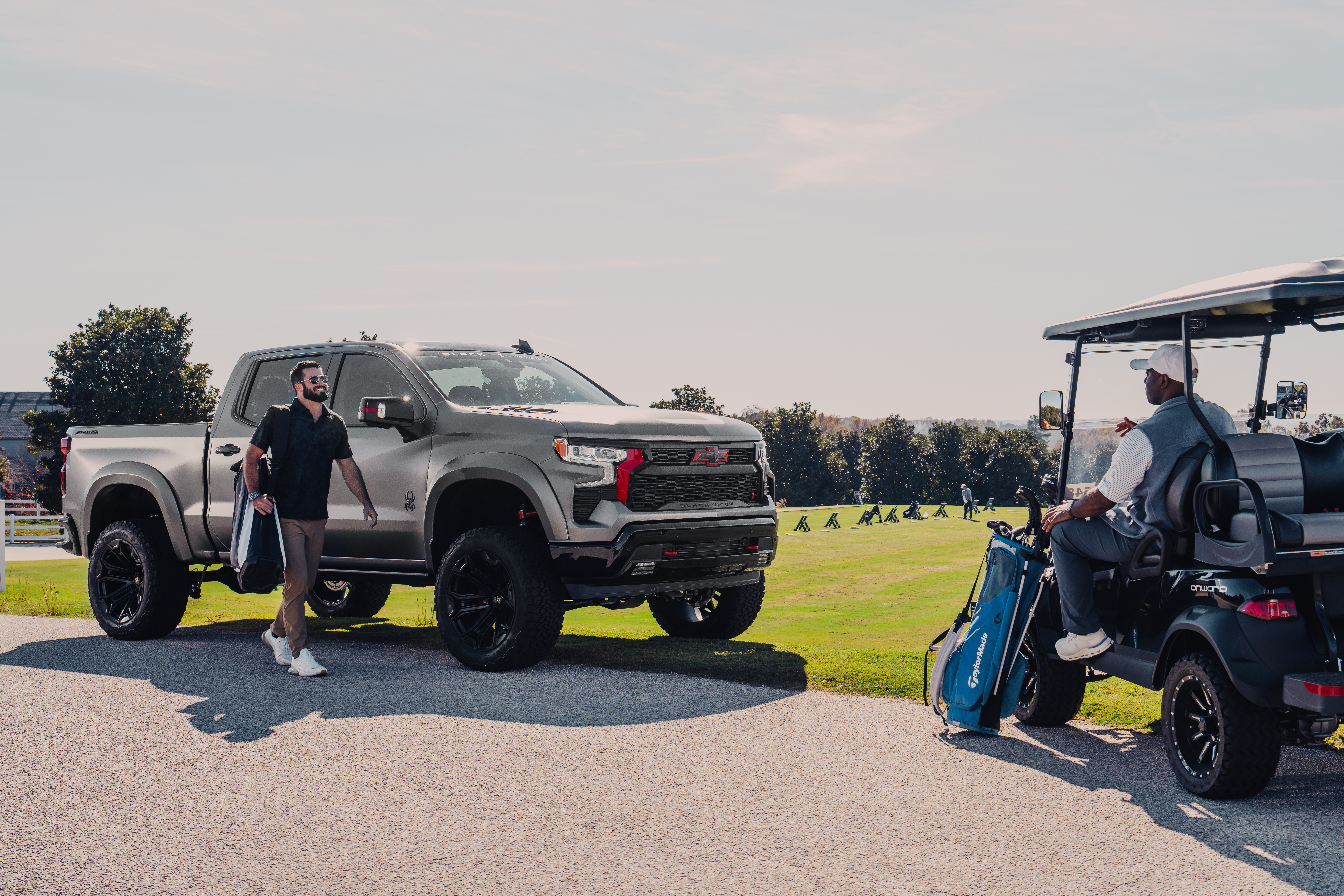 matte silver - grey Chevrolet Silverado hd 2500 black widow limited lifted truck with custom grille and red accents parked at a driving range with a man smiling carrying a golf bag walking towards a golf cart