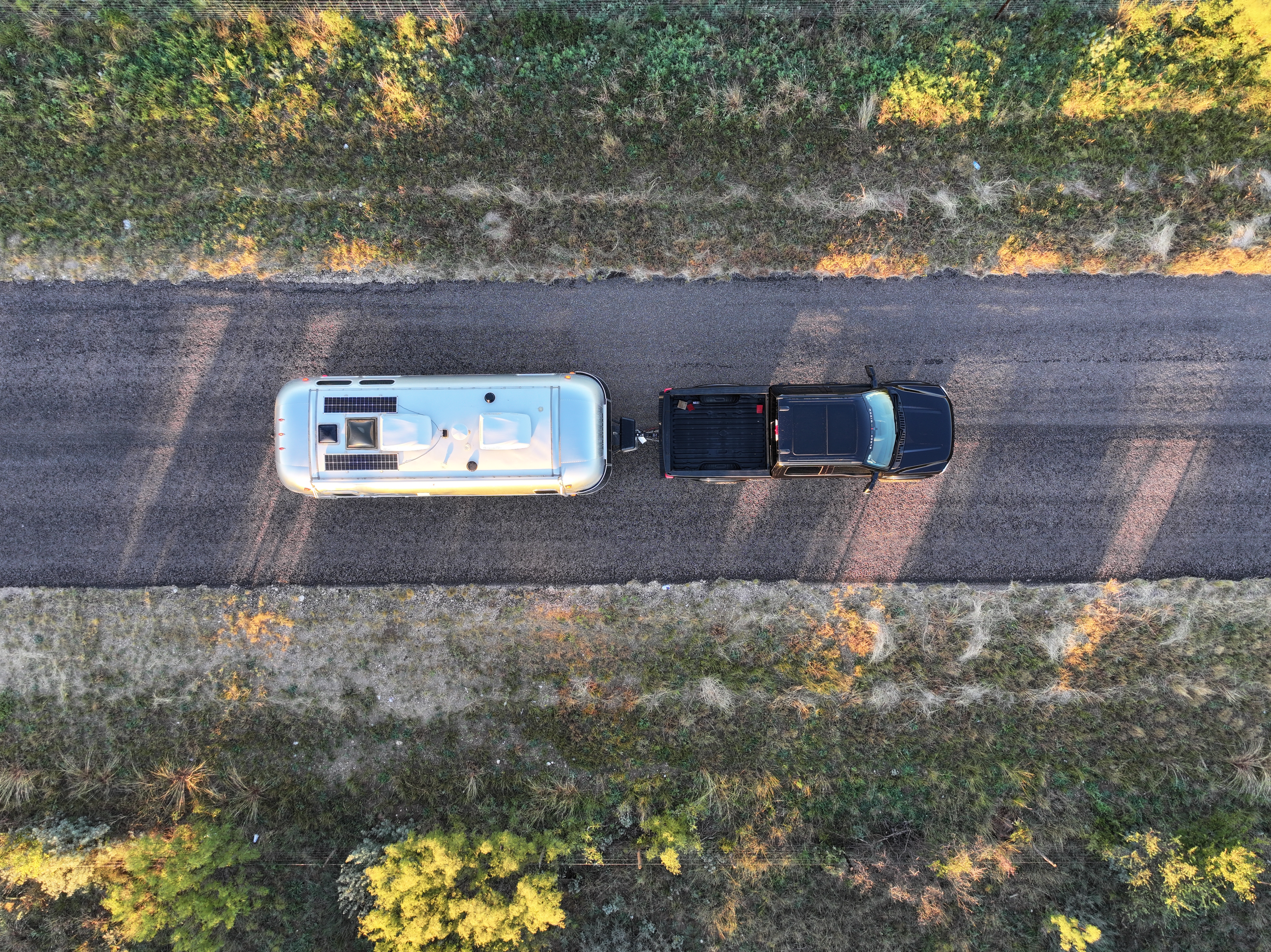 drone shot of a black ford black widow truck towing a small air stream on a two lane road 