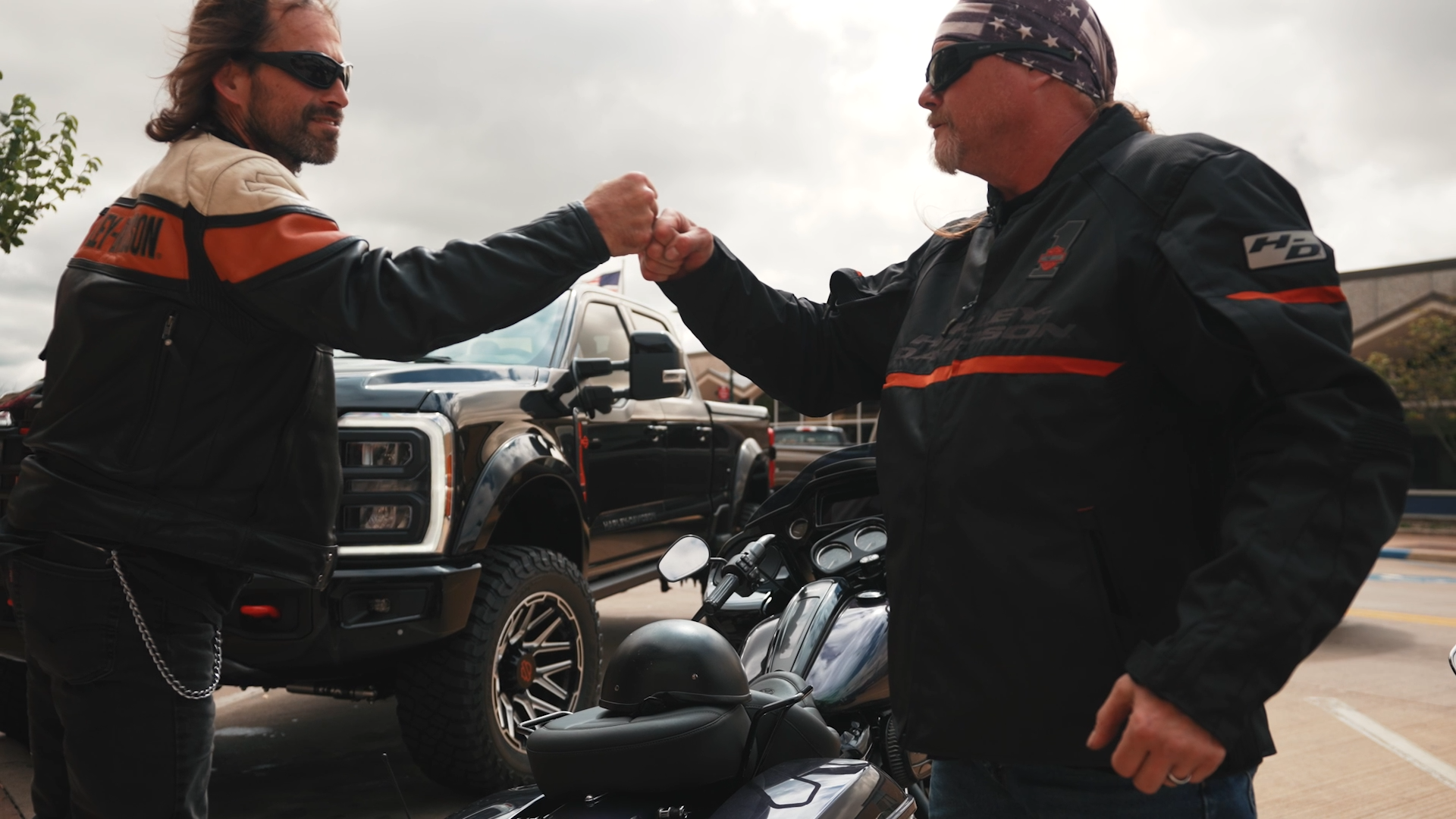 two motorycle riders fist bumping in front of a lifted black Harley-Davidson truck