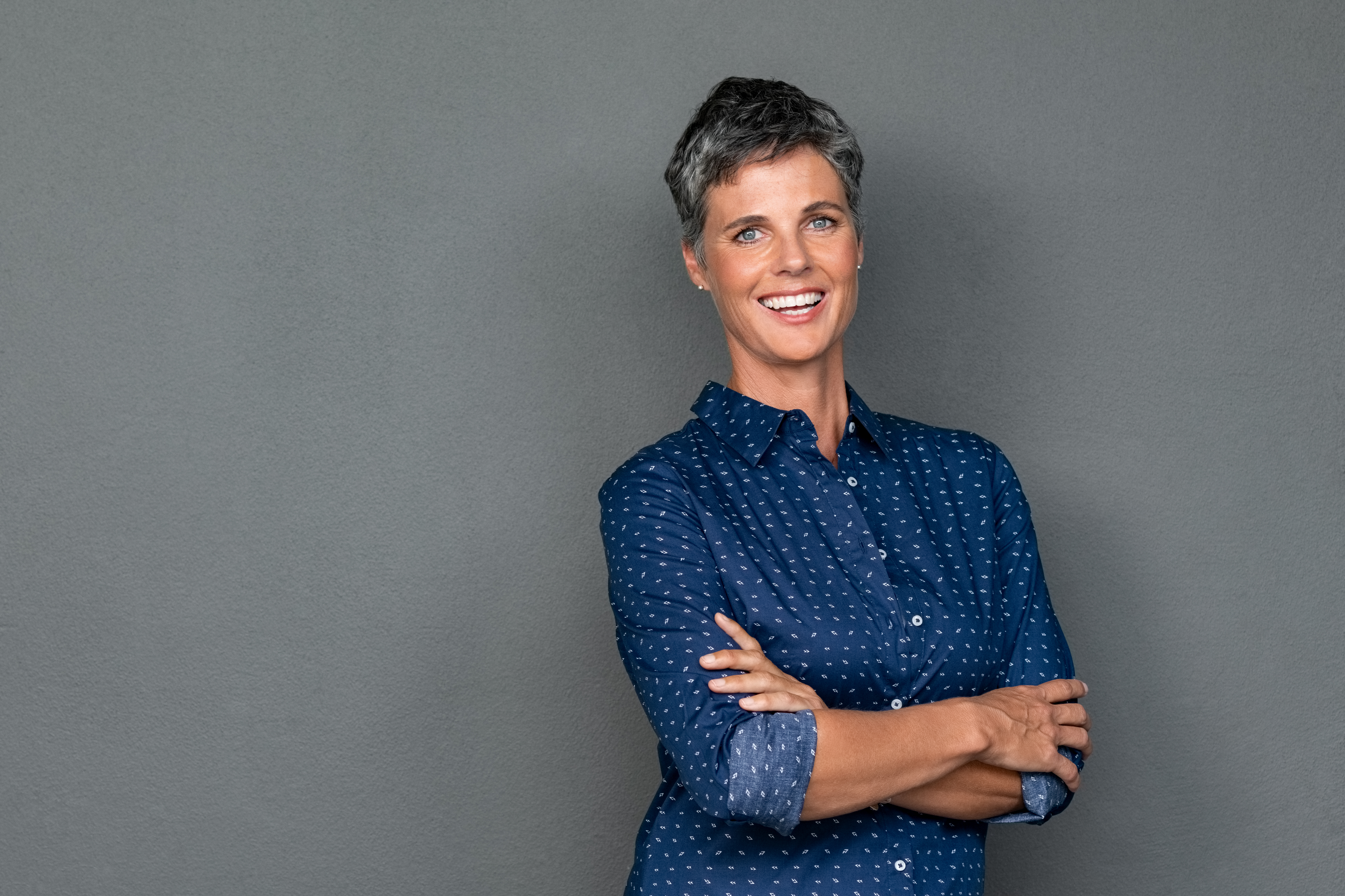 Isolated portrait of successful businesswoman with crossed arms looking at camera. Portrait of happy mature woman standing with arms folded isolated over gray background. Confident business woman smiling with copy space.